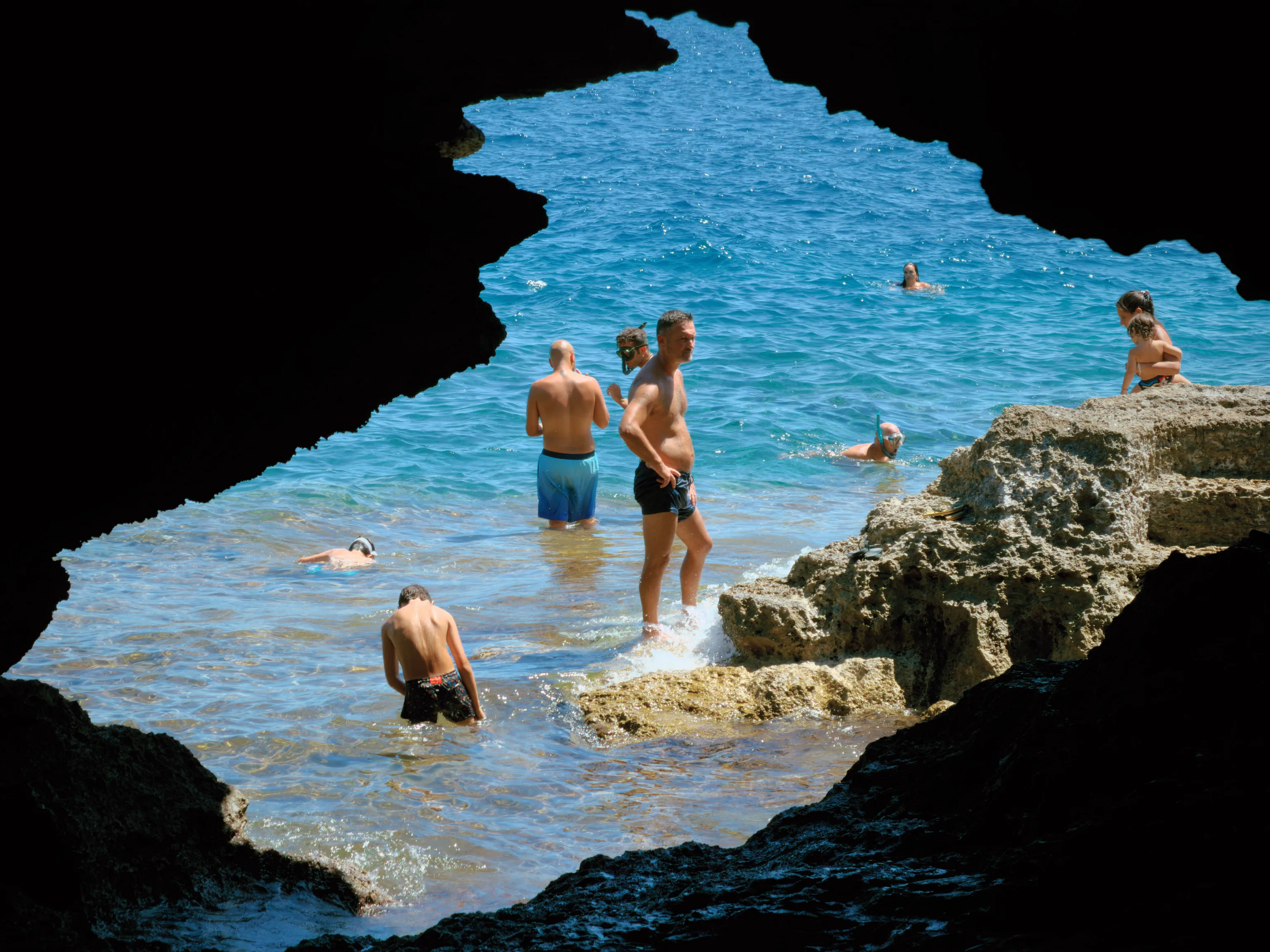Swimmers and waders stand in bright turquoise shallows below rocky ledges, framed by the jagged black silhouette of a sea cave opening in strong contrast to the sunlit water.