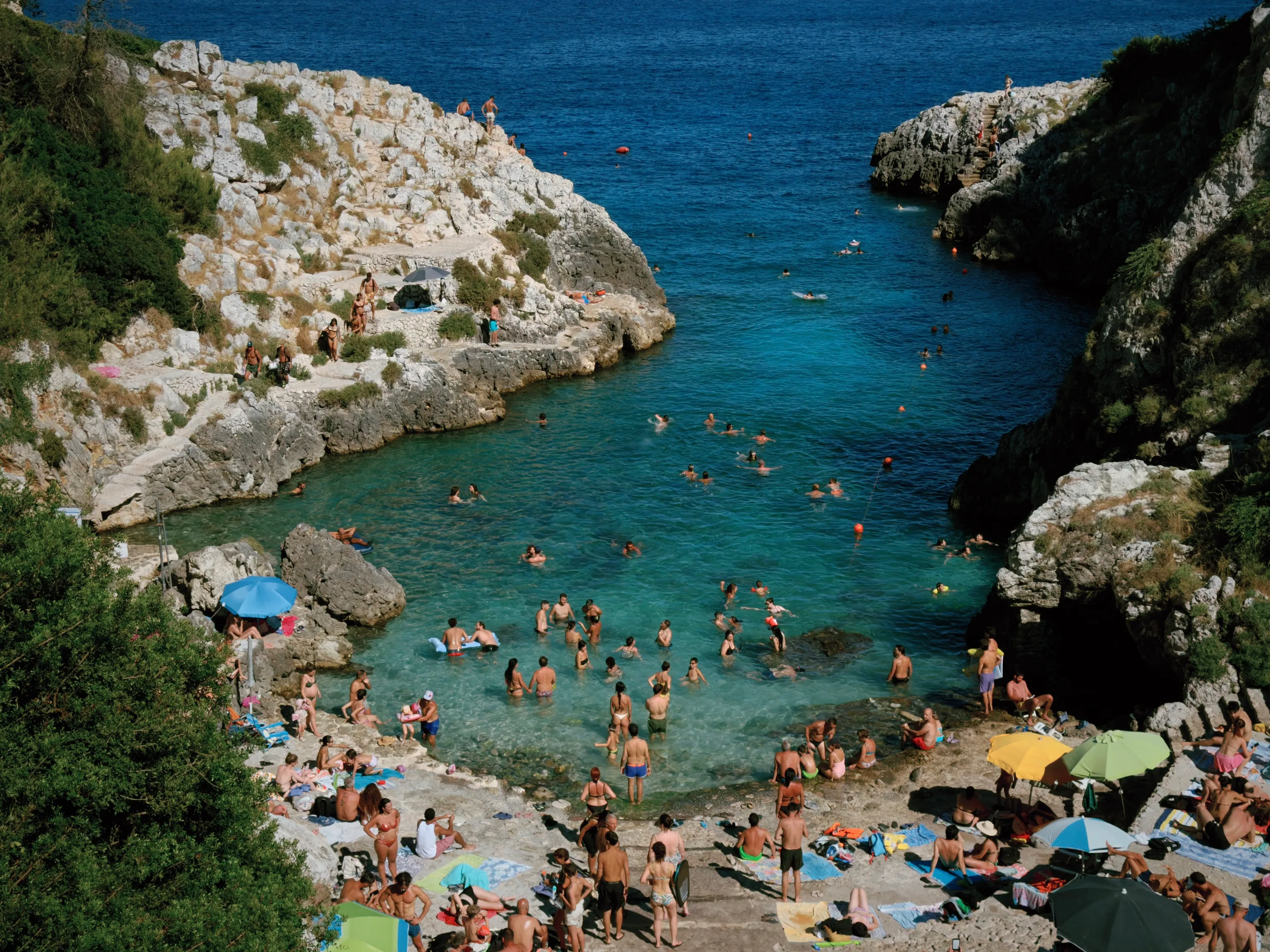 Sunbathers and swimmers crowd a rocky cove, its shallow turquoise water contrasting with the deep blue sea beyond sunlit limestone cliffs.