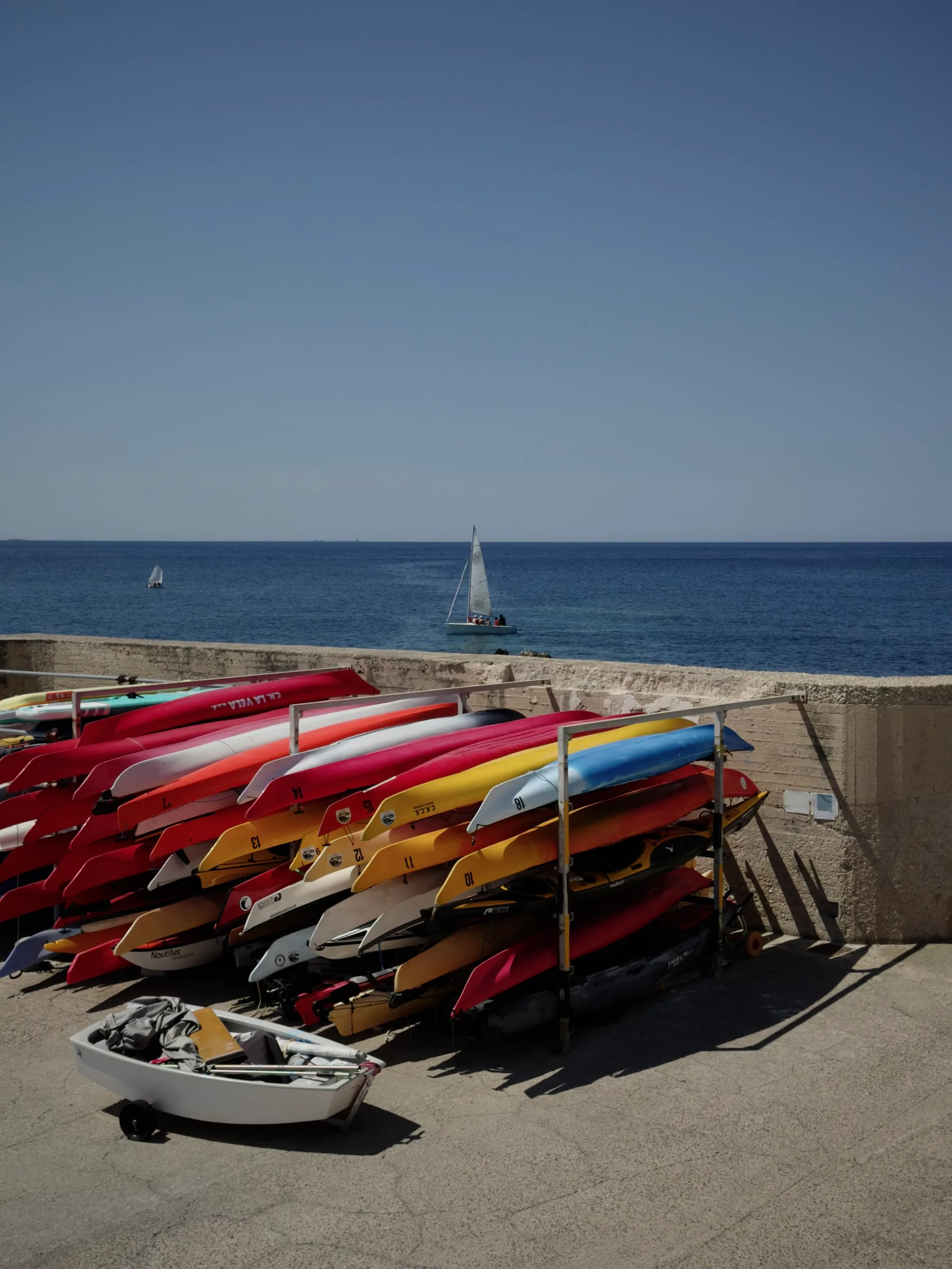 Colorful stacked kayaks in red, yellow, and blue line racks against a stone seawall while two sailboats glide across the deep blue sea beneath a clear sky.