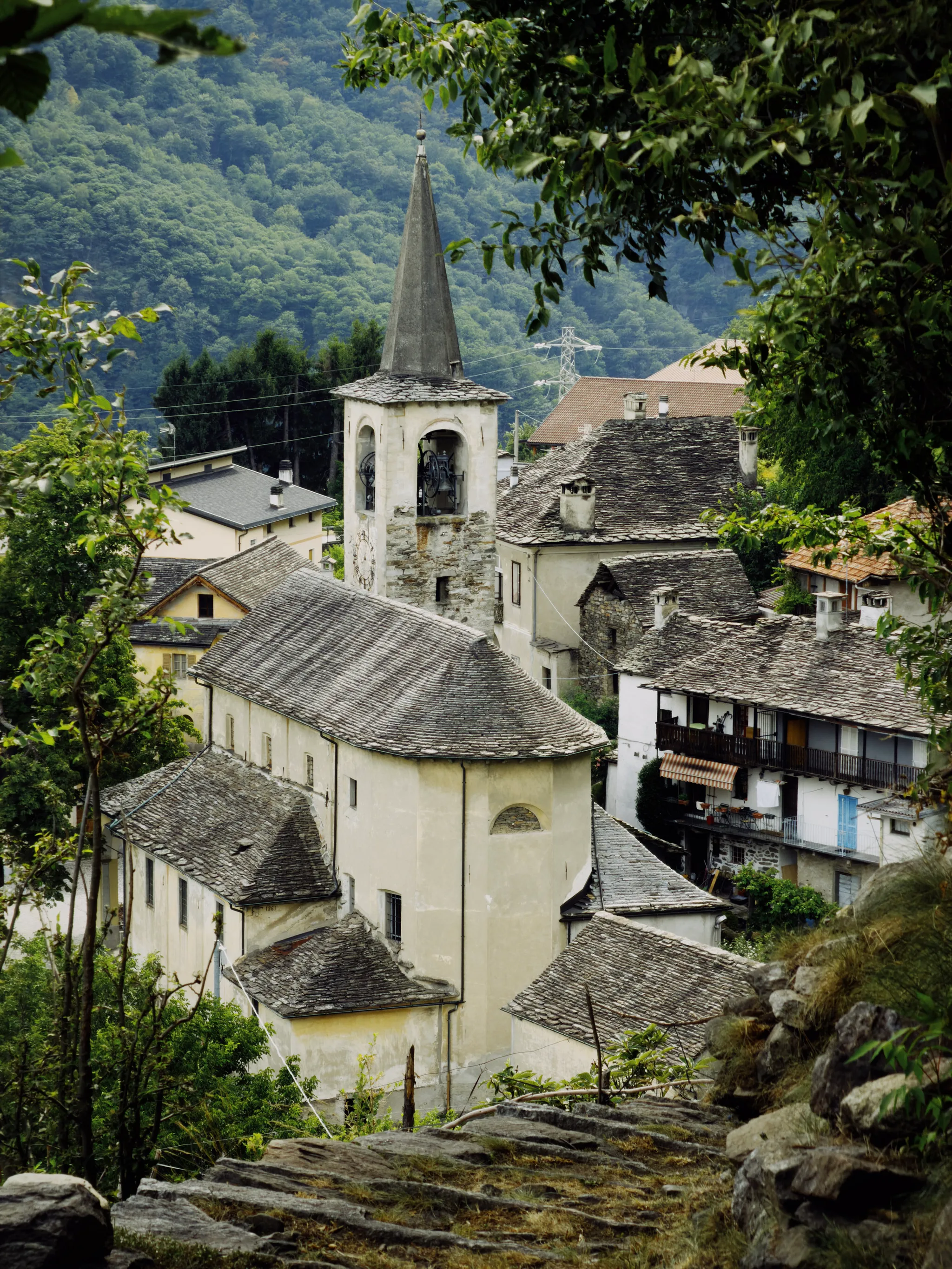 A cream-colored village church with a tall pointed bell tower rises among grey stone-roofed houses, framed by overhanging green leaves against a forested mountainside.