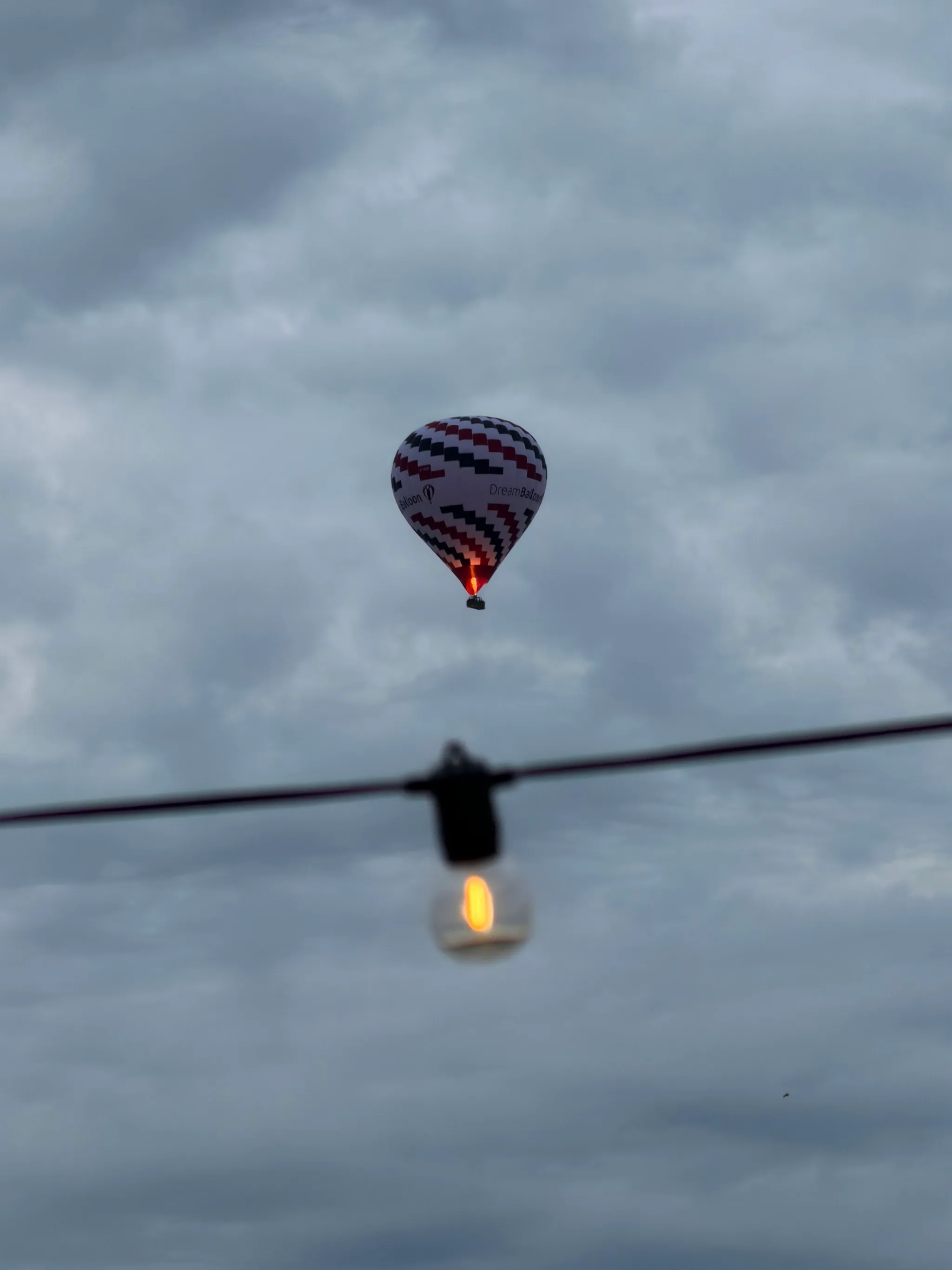 A red, white, and black patterned hot air balloon drifts against a gray overcast sky, a single glowing bulb on a string light hanging blurred in the foreground.