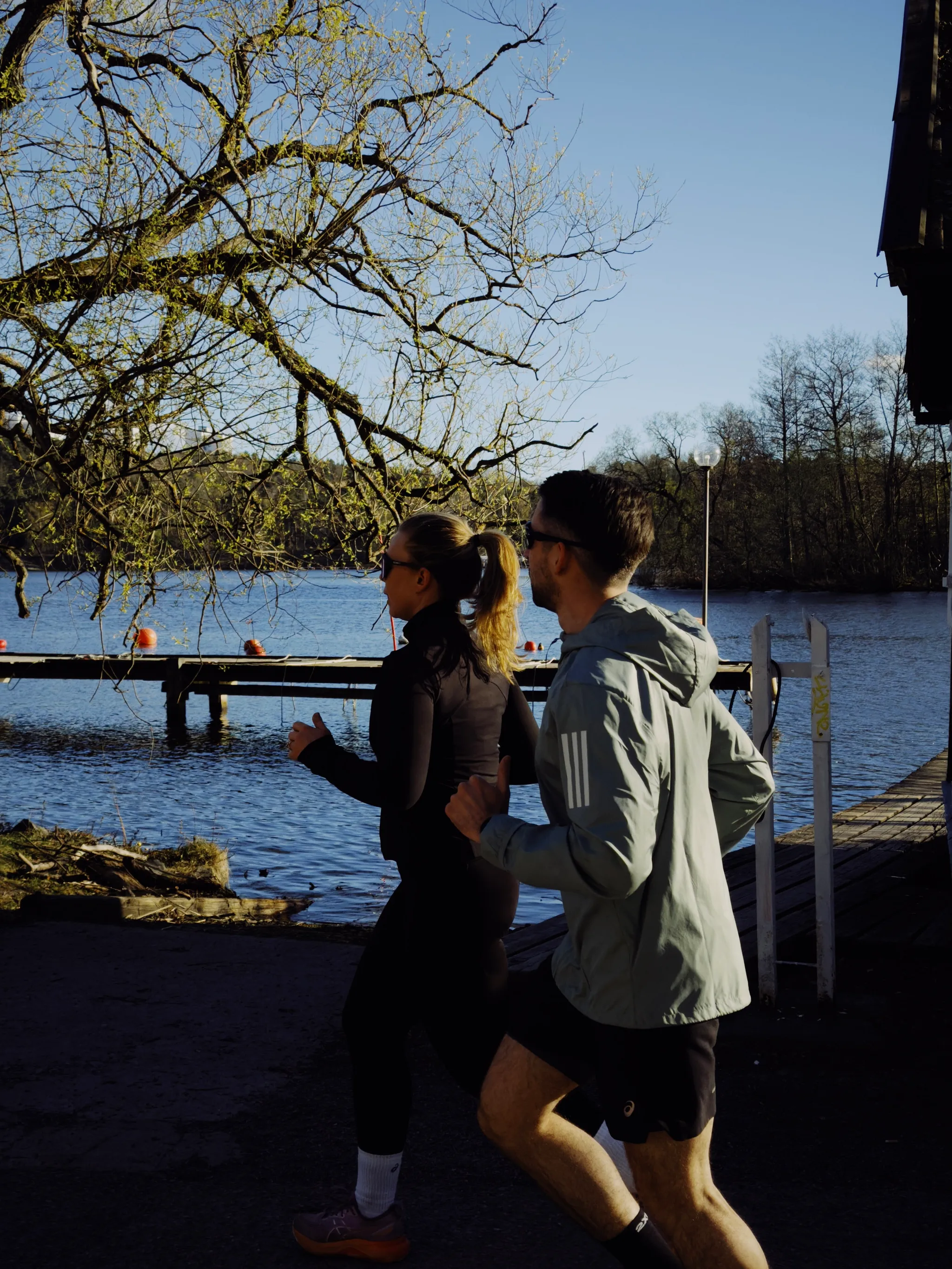Two joggers run past a wooden dock on a calm Stockholm lake beneath budding branches, fresh spring foliage catching bright sunlight against deep shadows.
