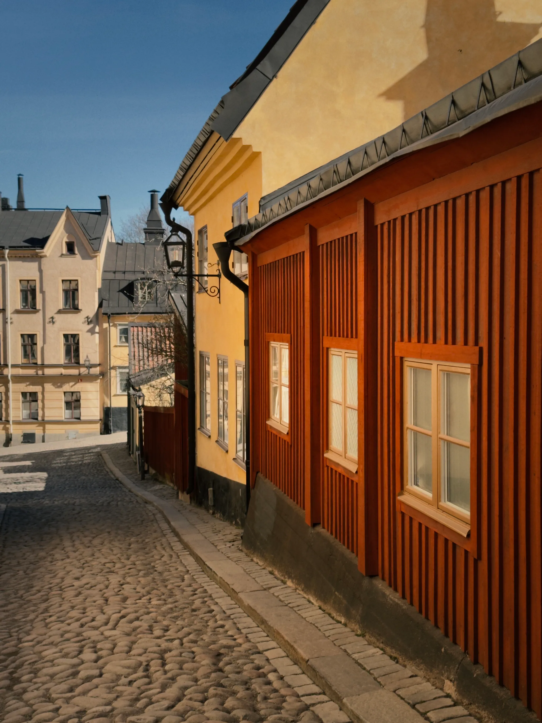 Sunlight warms a cobbled Stockholm street where an orange wooden building and ochre-yellow facades line the curve under a clear blue sky.