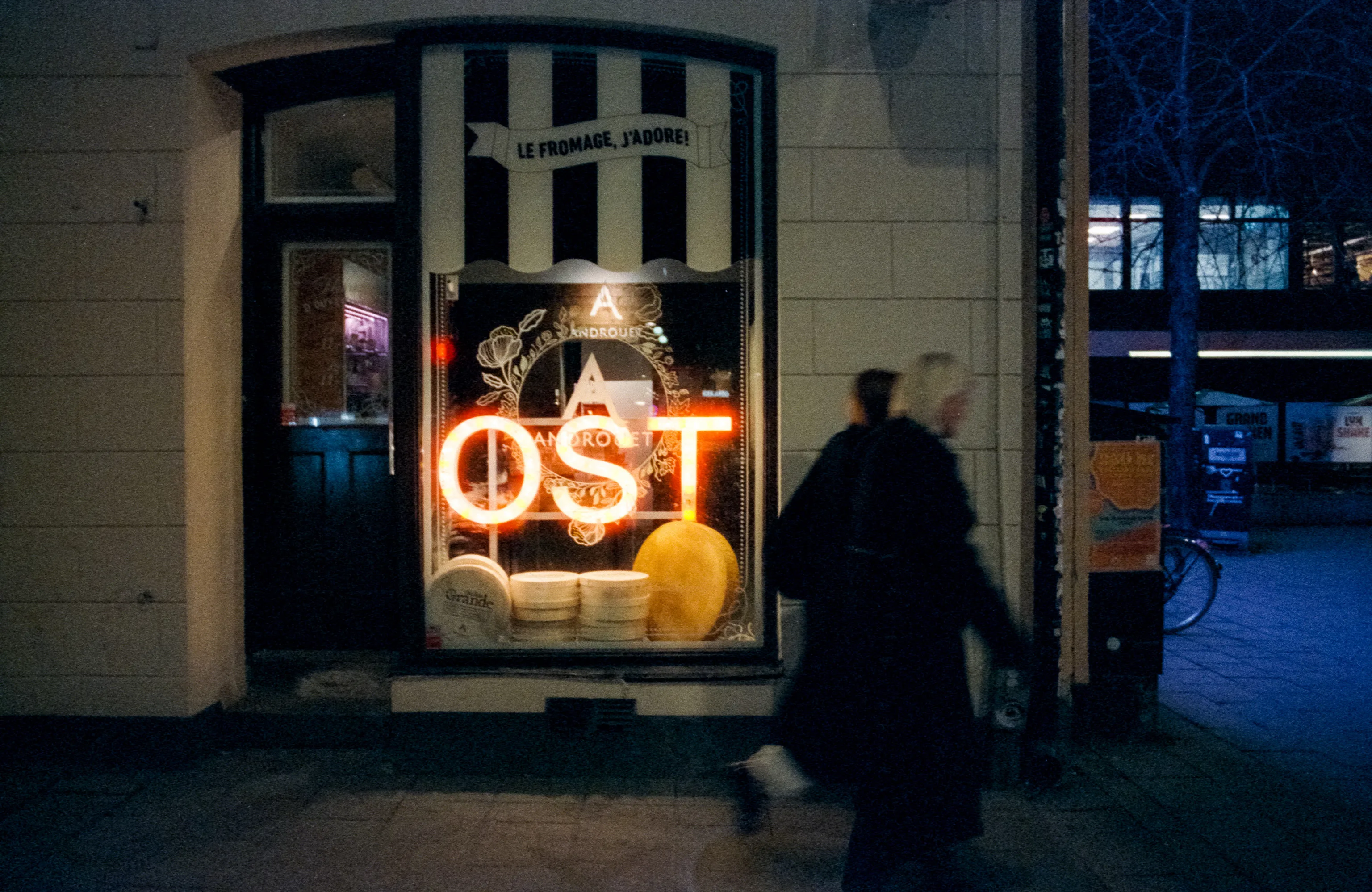 At night, a cheese shop's glowing orange neon sign reading OST illuminates two motion-blurred pedestrians walking past, the warm light contrasting with cool blue street tones.