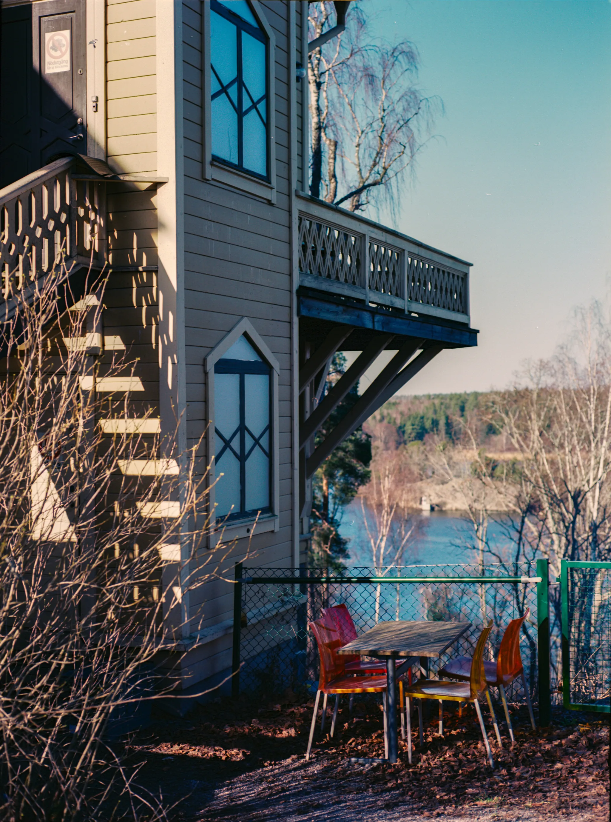 Sunlit wooden house with a carved balcony above a table and bright orange chairs by a fence, bare branches framing a blue lake and shoreline under a clear sky.