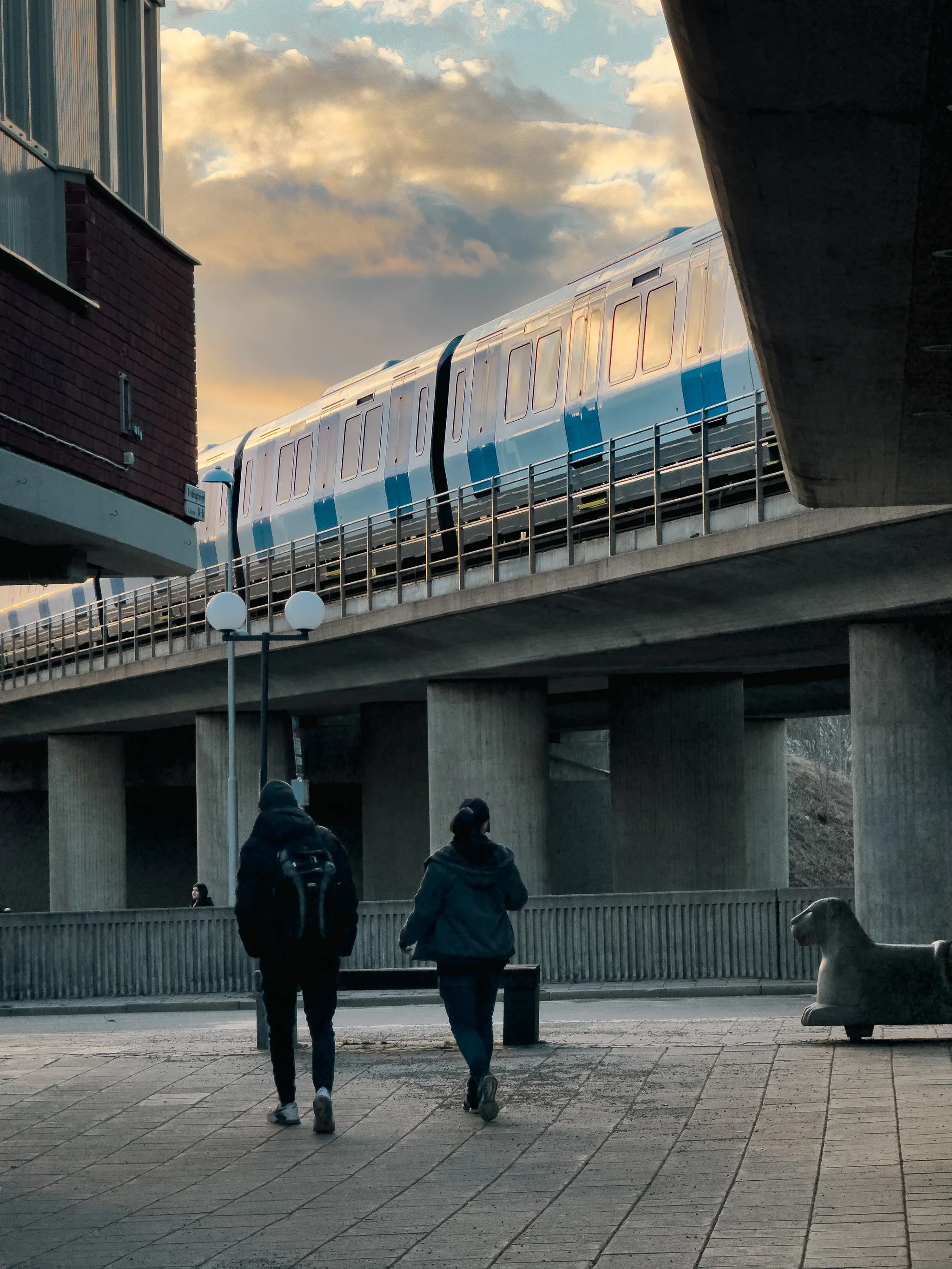 Two people walk across a paved plaza beneath a concrete viaduct in Stockholm as a blue metro train passes overhead, its windows catching warm golden sunset light under glowing clouds.