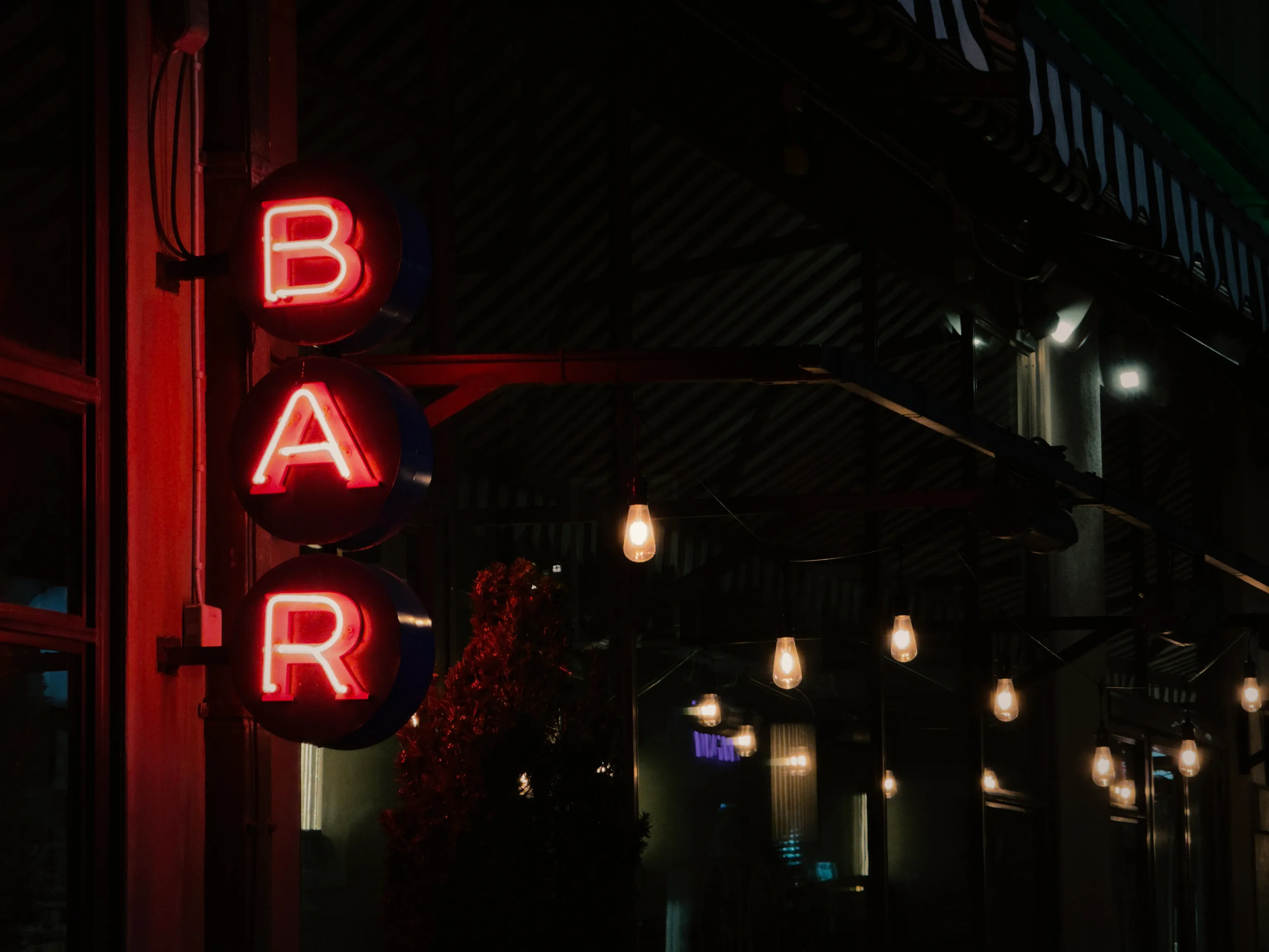 Three vertical red neon letters spelling BAR glow against a dark building at night, with warm Edison bulbs hanging beneath an awning in the dim background.