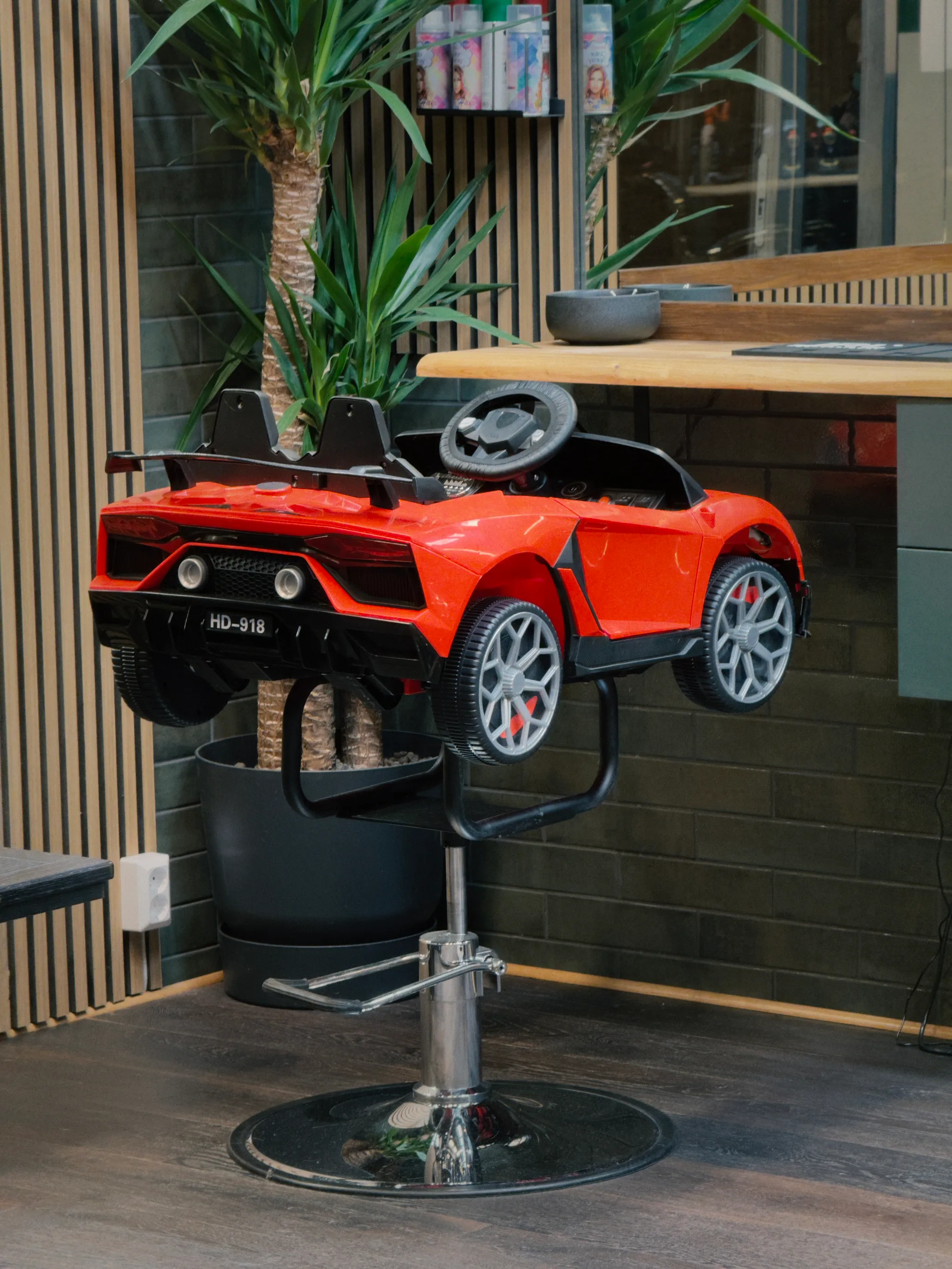 In a barbershop, a red toy sports car sits mounted on a hydraulic chair base, its glossy scarlet paint standing out against dark green tiles and a tall potted palm.