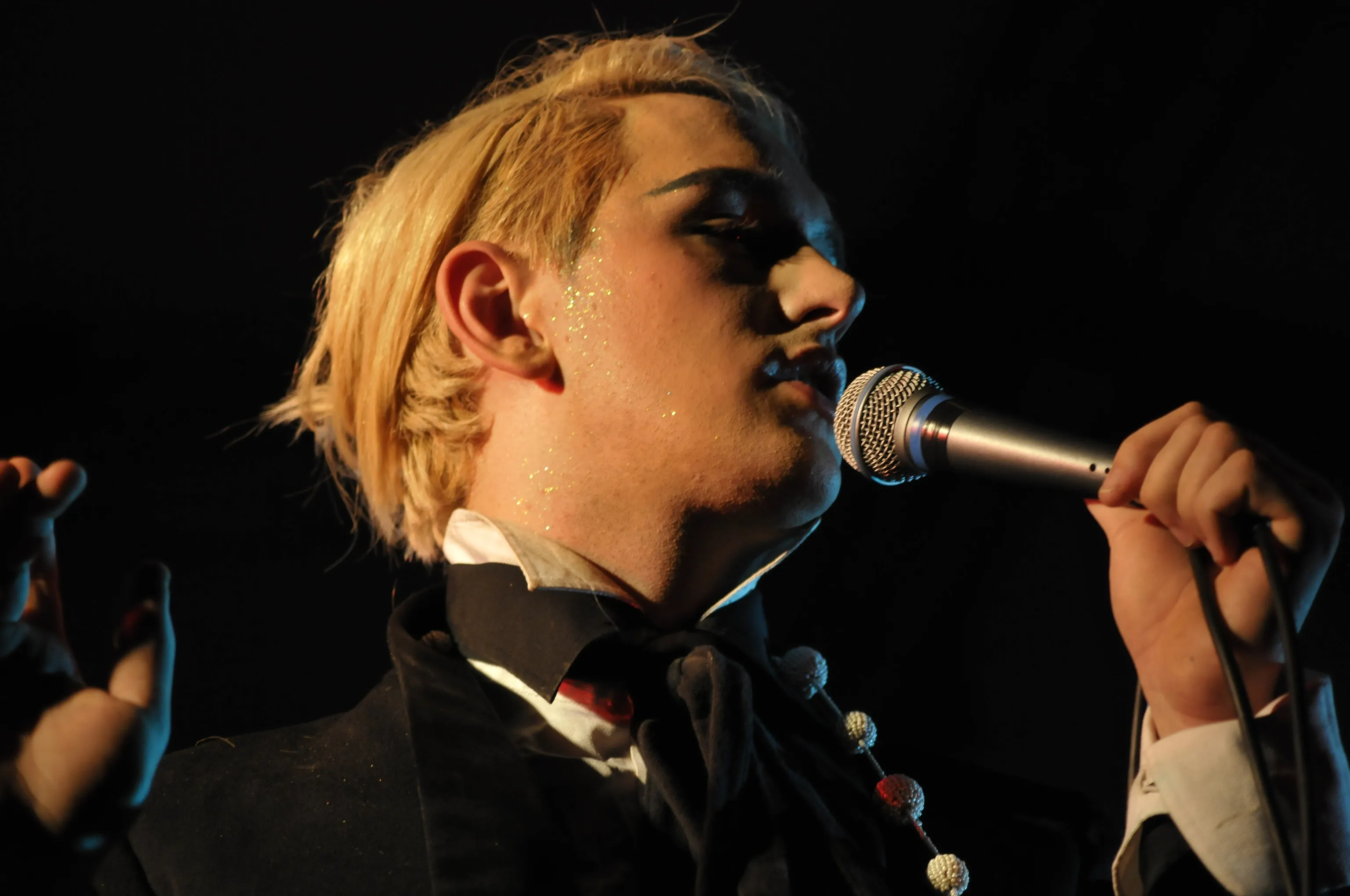 Blonde singer with glitter dusted on his cheek sings into a microphone, eyes closed in profile, lit by warm stage light against a black background.