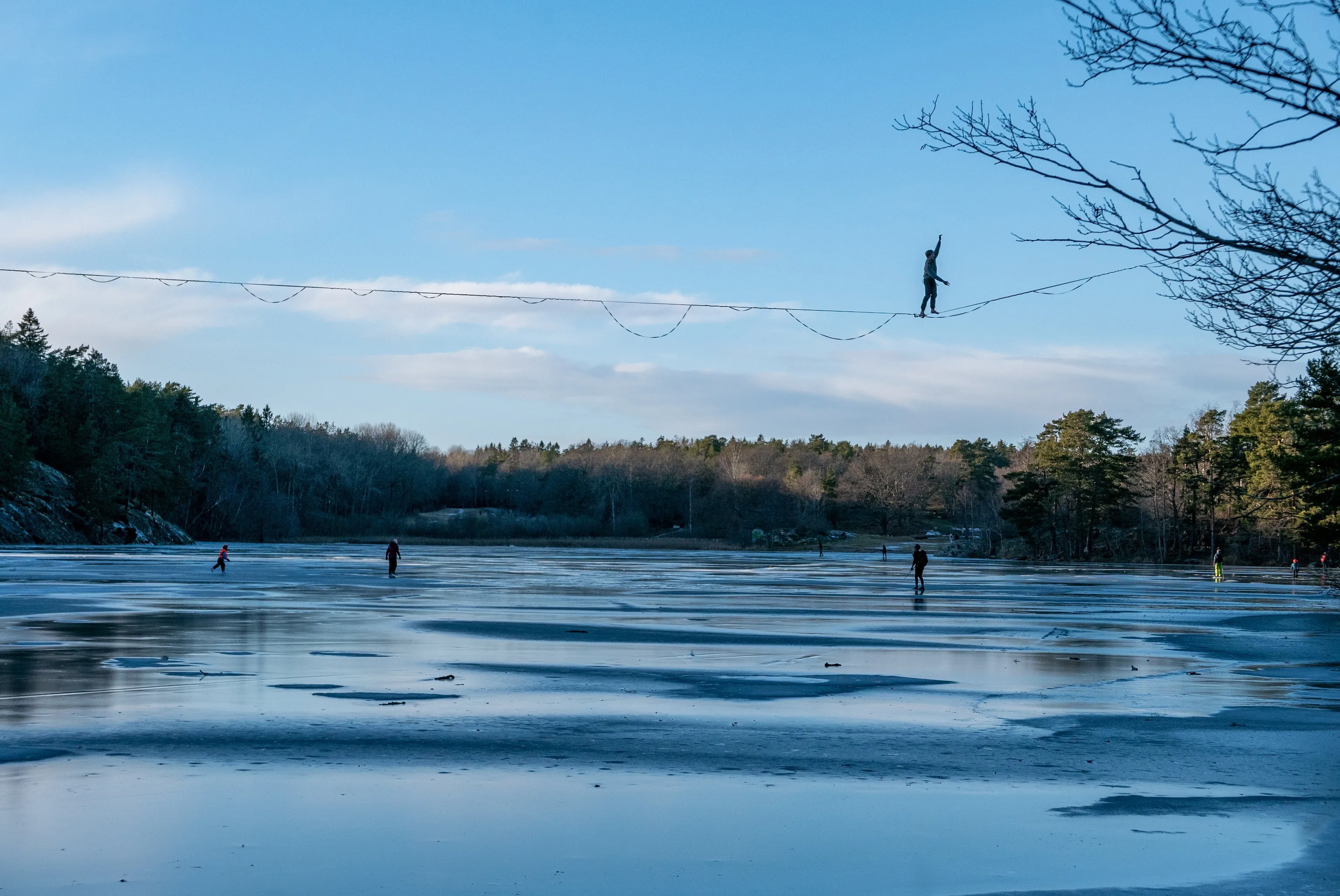 A slackliner balances on a line strung high above a frozen lake while skaters glide across the blue ice below, silhouetted against a pale winter sky framed by bare branches.
