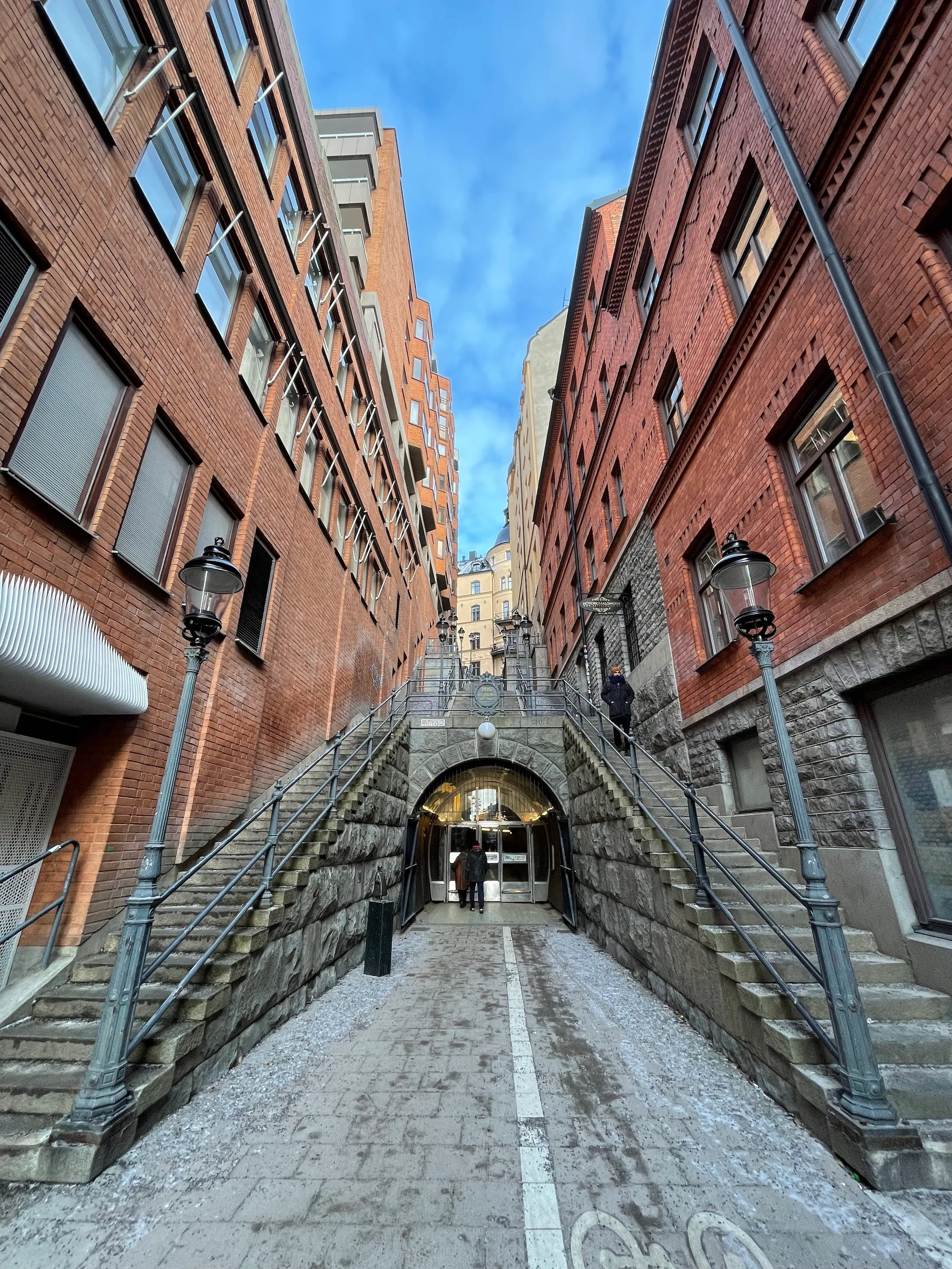 Symmetrical stone staircases rise between tall red brick buildings toward an arched tunnel entrance, framed by ornate black lampposts under a bright blue sky.