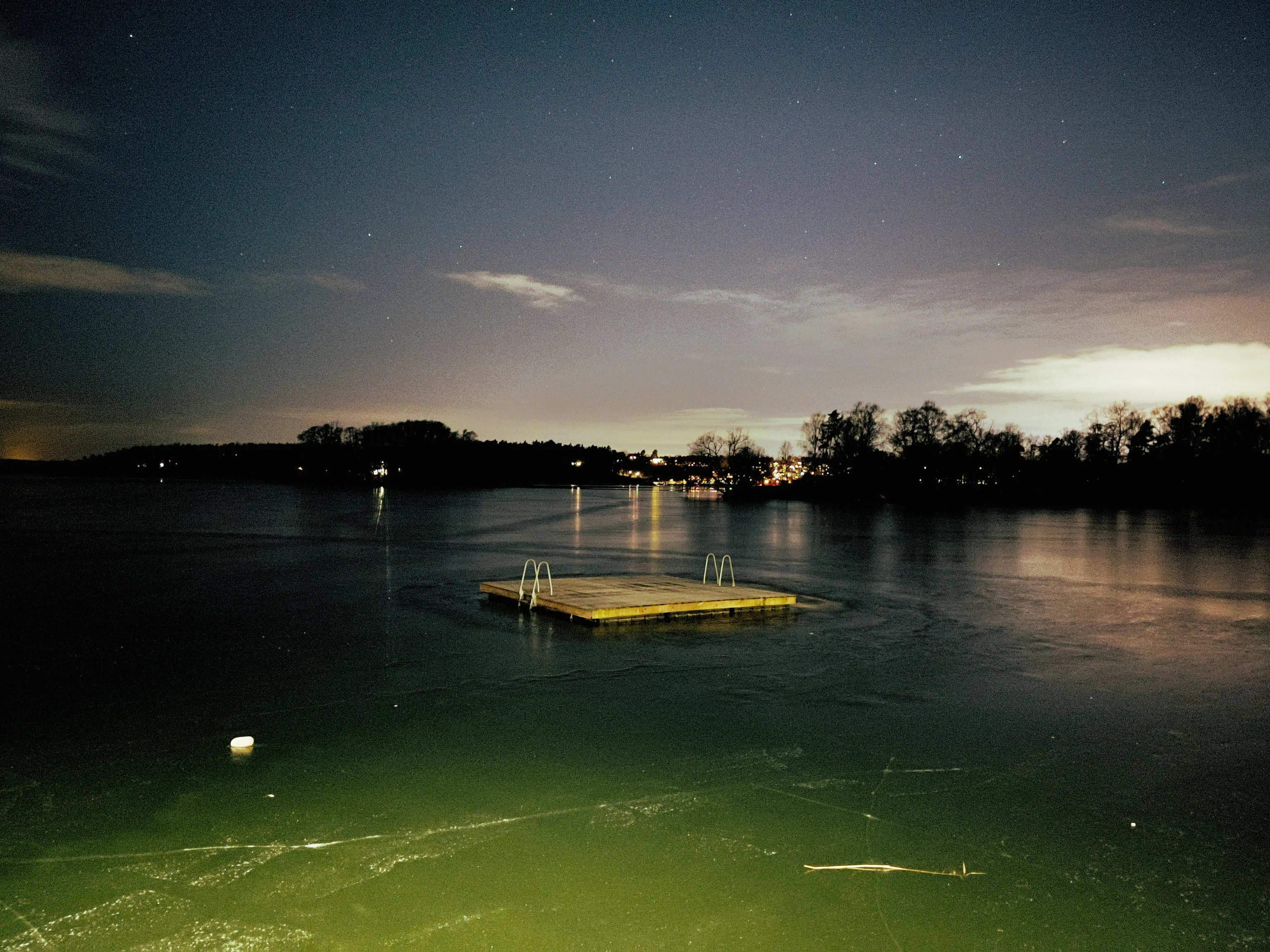 A wooden swim platform with two metal ladders sits on partially frozen dark green water beneath a starry night sky, with silhouetted trees and distant shoreline lights glowing on the horizon.