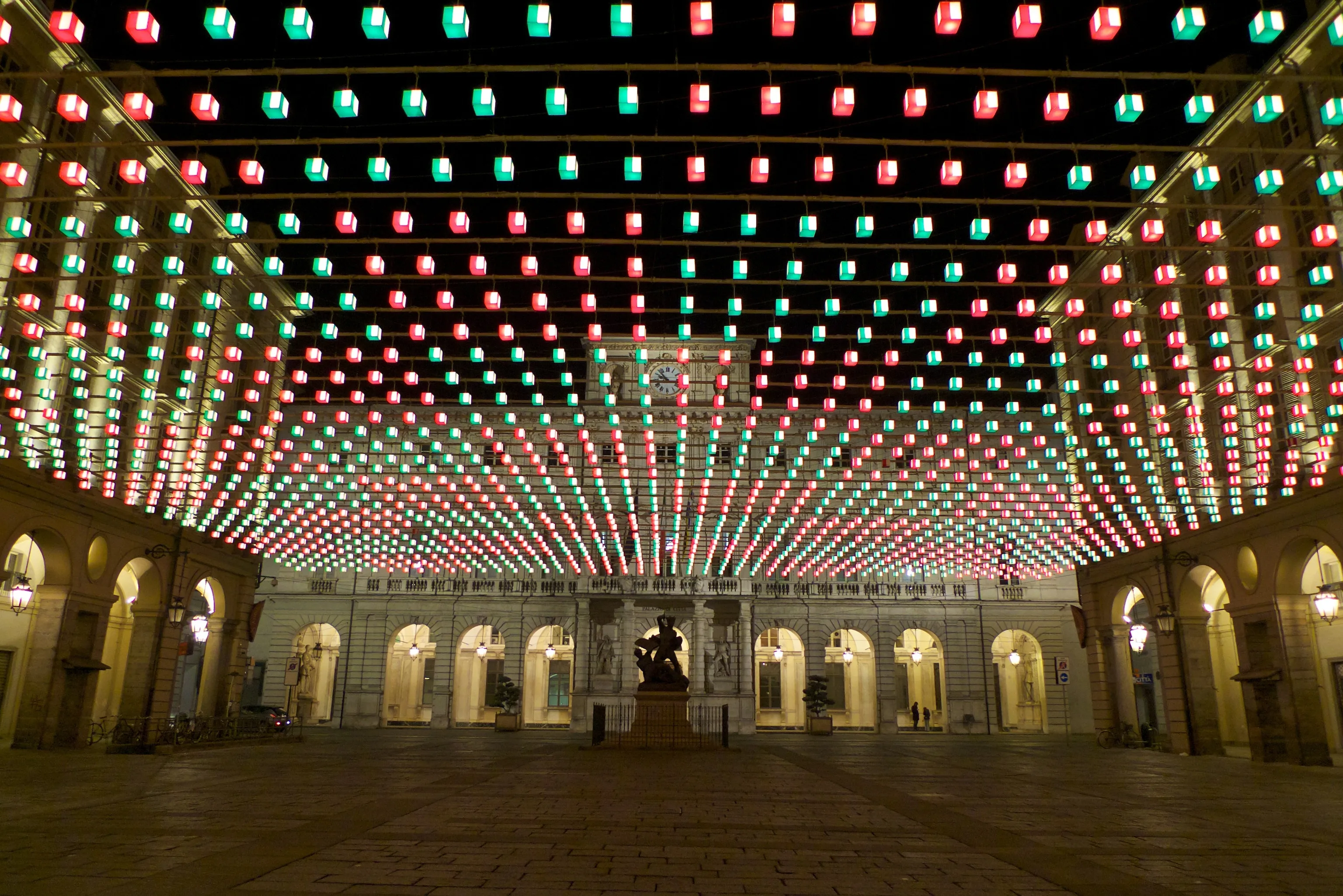 Rows of glowing red and green lanterns strung overhead converge above an empty paved square toward an ornate floodlit building facade at night, with an equestrian statue silhouetted at center.