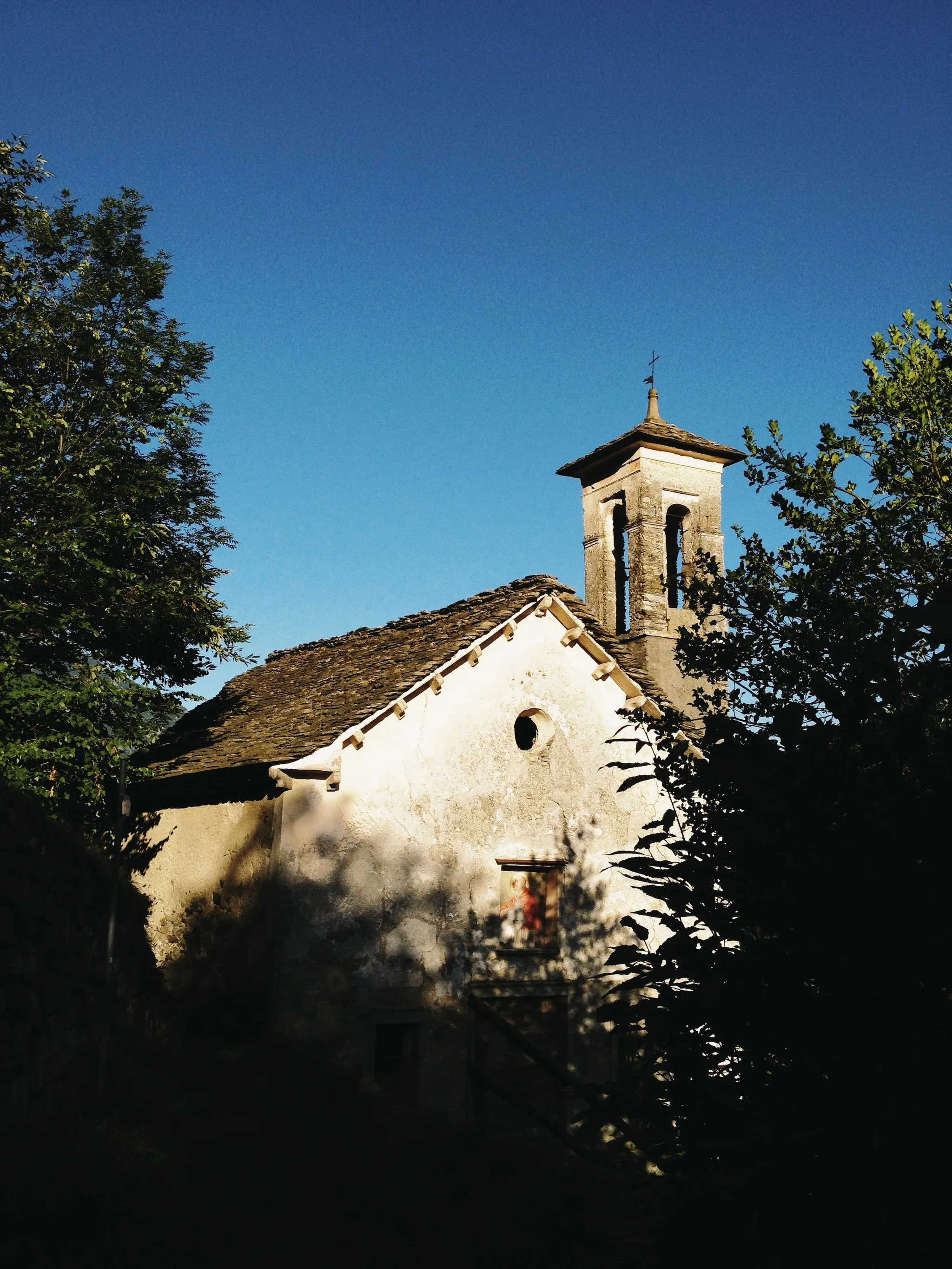An old whitewashed stone chapel with a small bell tower peeks through dark trees under a deep blue sky, its sunlit facade dappled with leaf shadows.