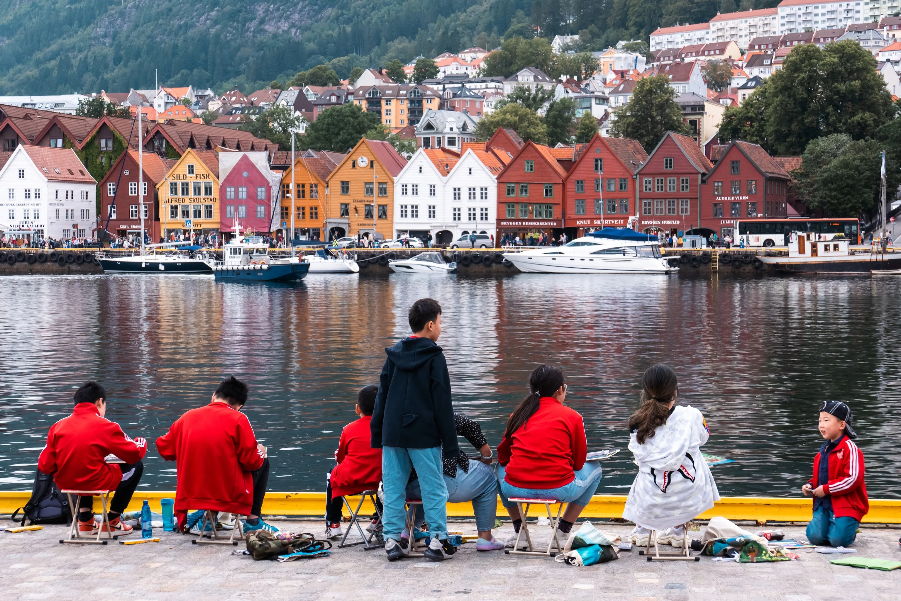 Children in matching red jackets sit on folding stools at a harbor's edge, painting a row of vividly colored gabled buildings whose forms reflect in the calm water.