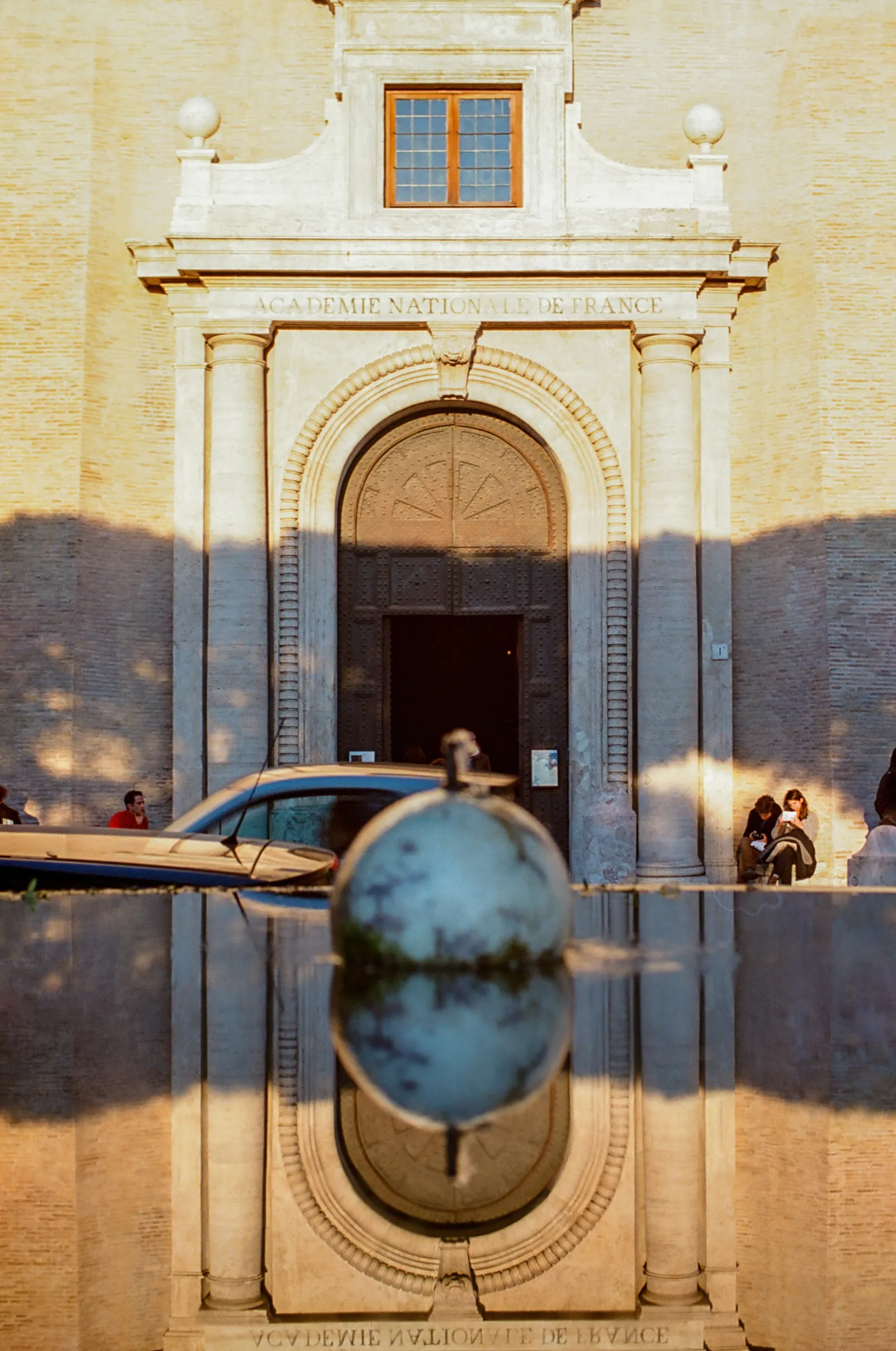 A stone sphere crowns a fountain before the Académie Nationale de France, whose warm-lit facade and arched doorway are mirrored almost perfectly in the still water below.