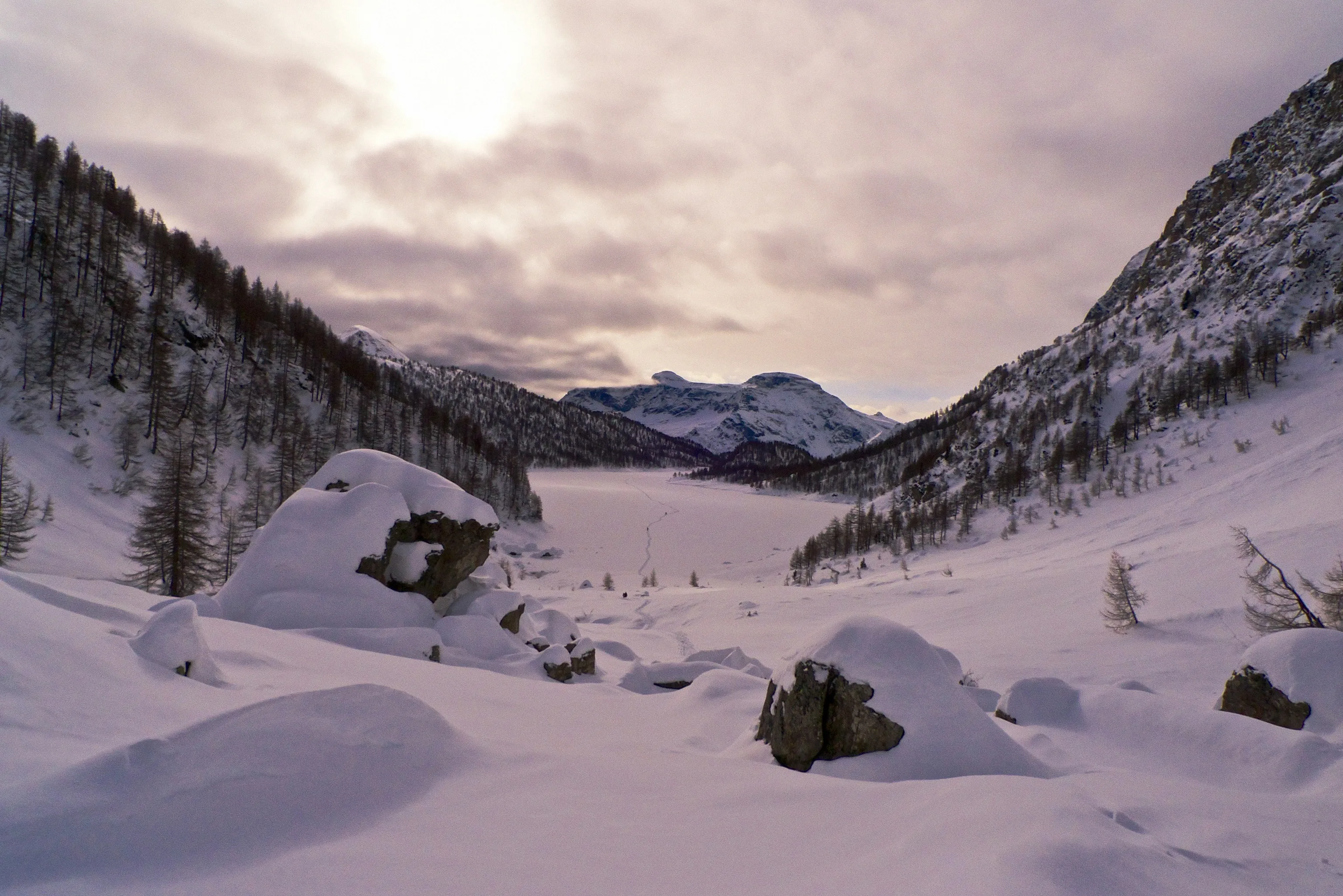 Snow-buried boulders overlook a frozen alpine lake ringed by evergreen slopes and snowy peaks, under thick clouds glowing soft pink where the sun breaks through.