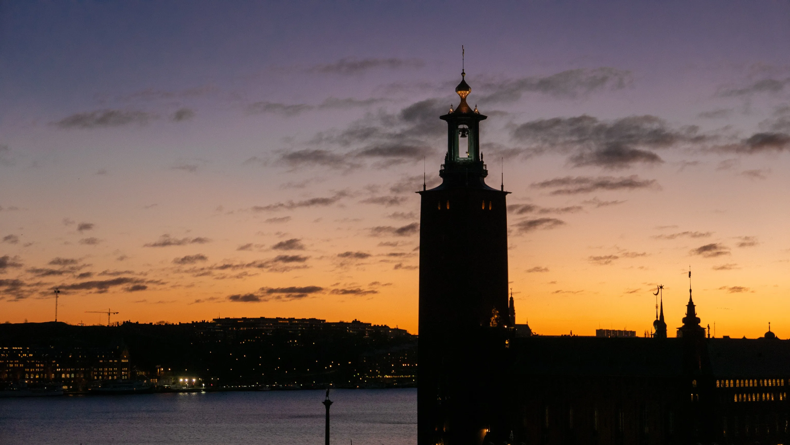 Stockholm City Hall's tower stands in dark silhouette over the water at dusk, its spire outlined against a violet-to-orange sunset sky with city lights glowing along the far shore.
