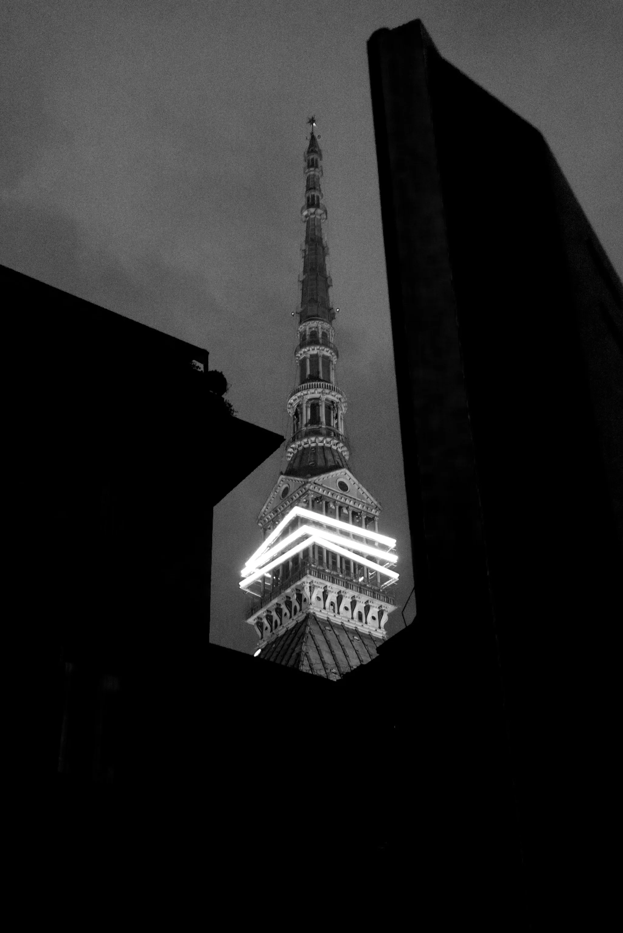 The illuminated Mole Antonelliana spire glows between black silhouettes of surrounding buildings at night, its bright lights a stark focal point in this grainy black-and-white composition.