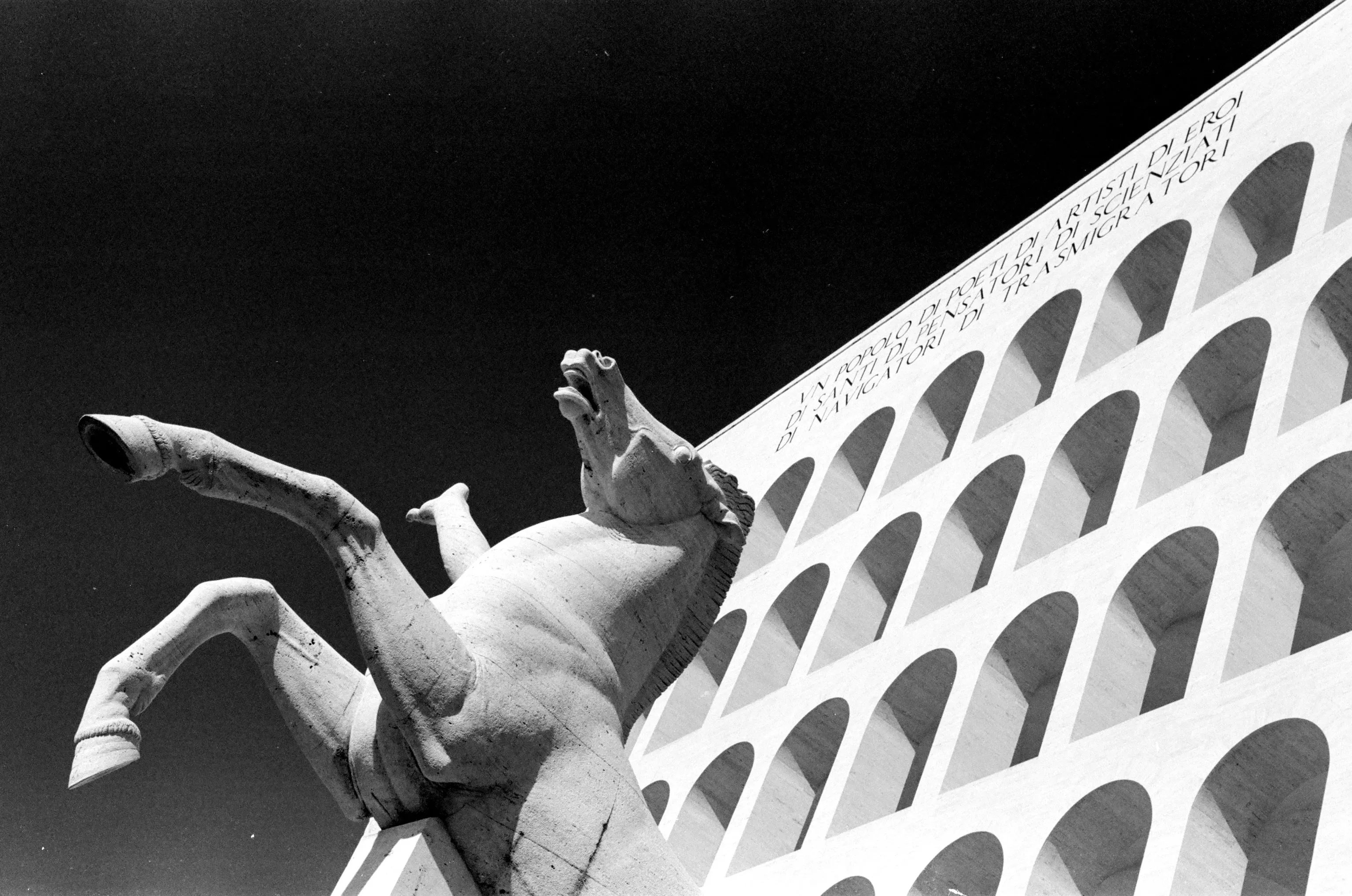A rearing stone horse sculpture looms against a deep black sky before the arch-filled facade of Rome's Square Colosseum, captured in stark black and white from a low angle.