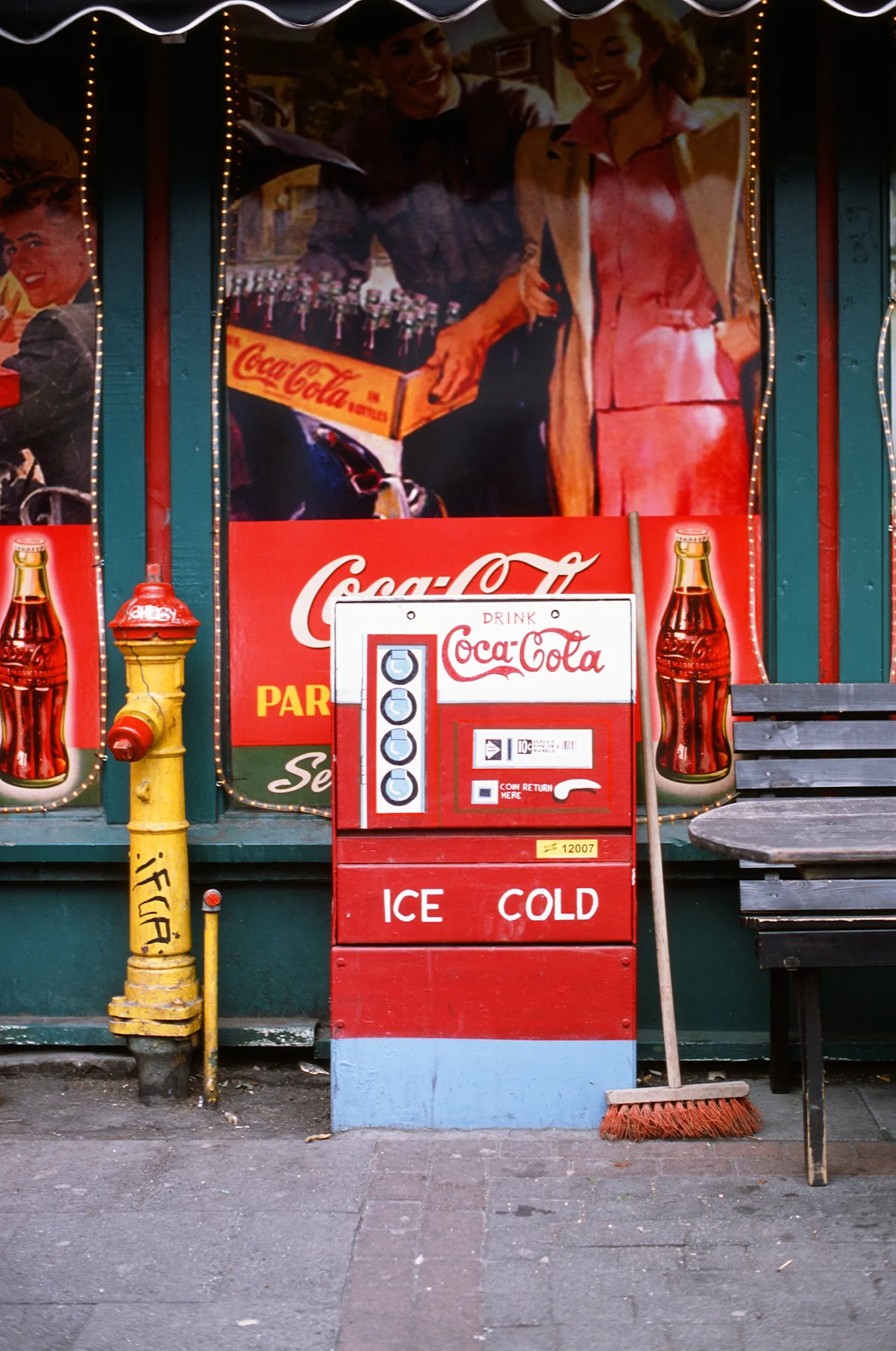 A red Coca-Cola machine stands on a sidewalk beside a yellow fire hydrant and broom, backed by a dark green storefront covered in vintage Coke murals glowing with rich, saturated color.