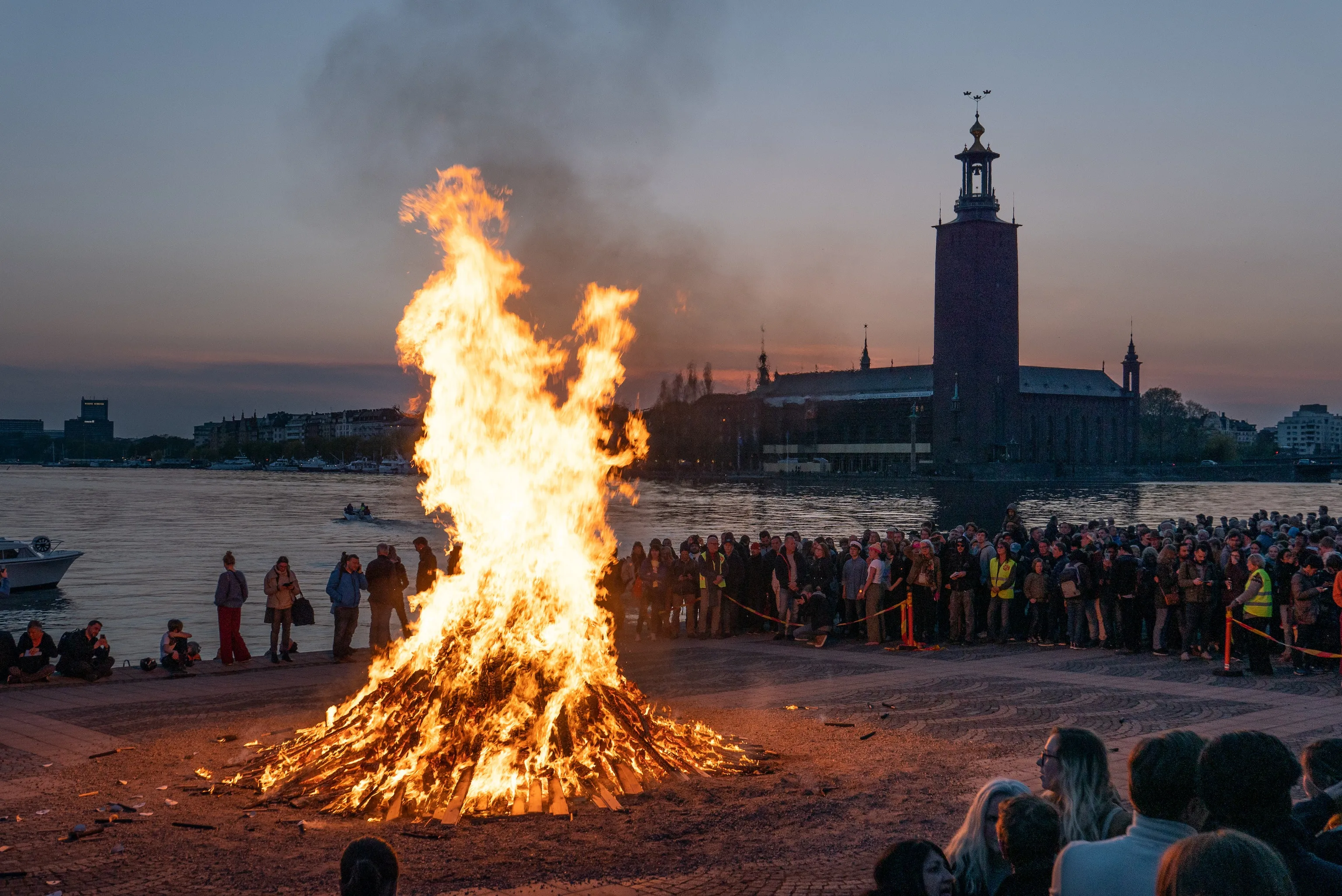 A huge bonfire blazes orange beside a waterfront crowd at dusk, Stockholm City Hall rising across the water under a pale twilight sky.