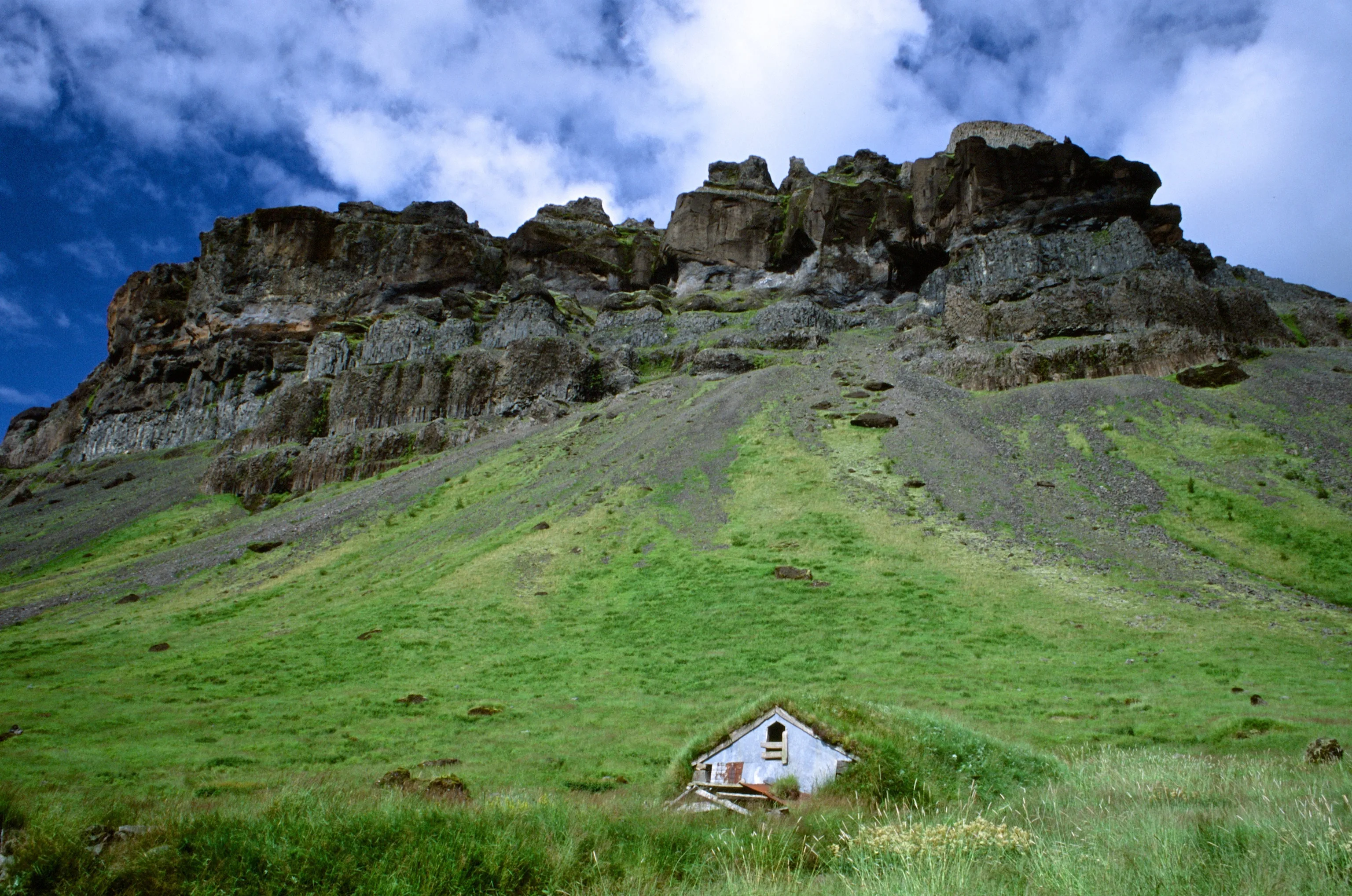 A small white turf-roofed house sits dwarfed on a vivid green slope beneath towering dark rock cliffs under a deep blue sky streaked with clouds.