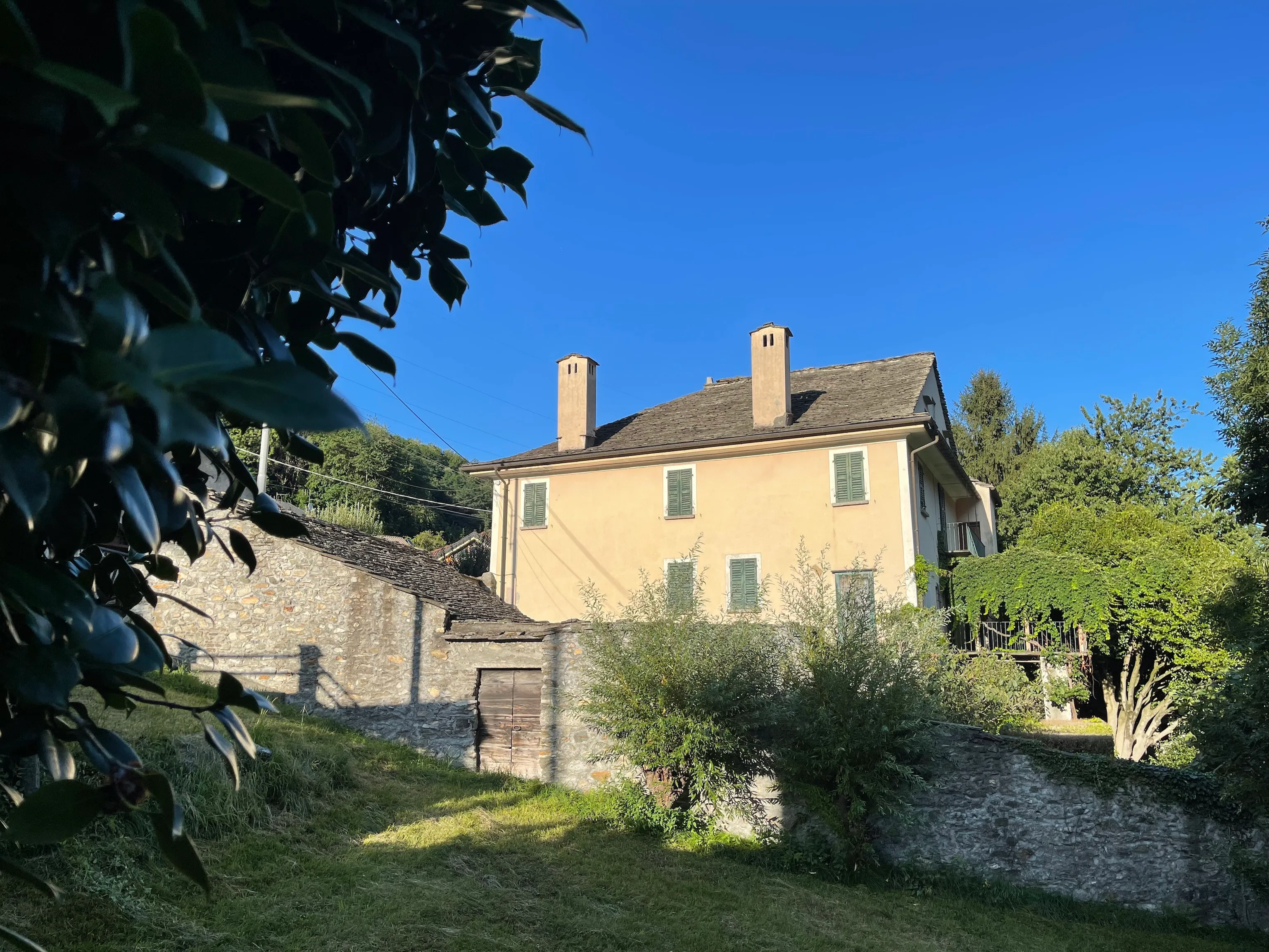 A peach stucco house with green shutters and twin chimneys sits behind old stone walls in warm evening light, framed by dark leaves against a deep blue sky.