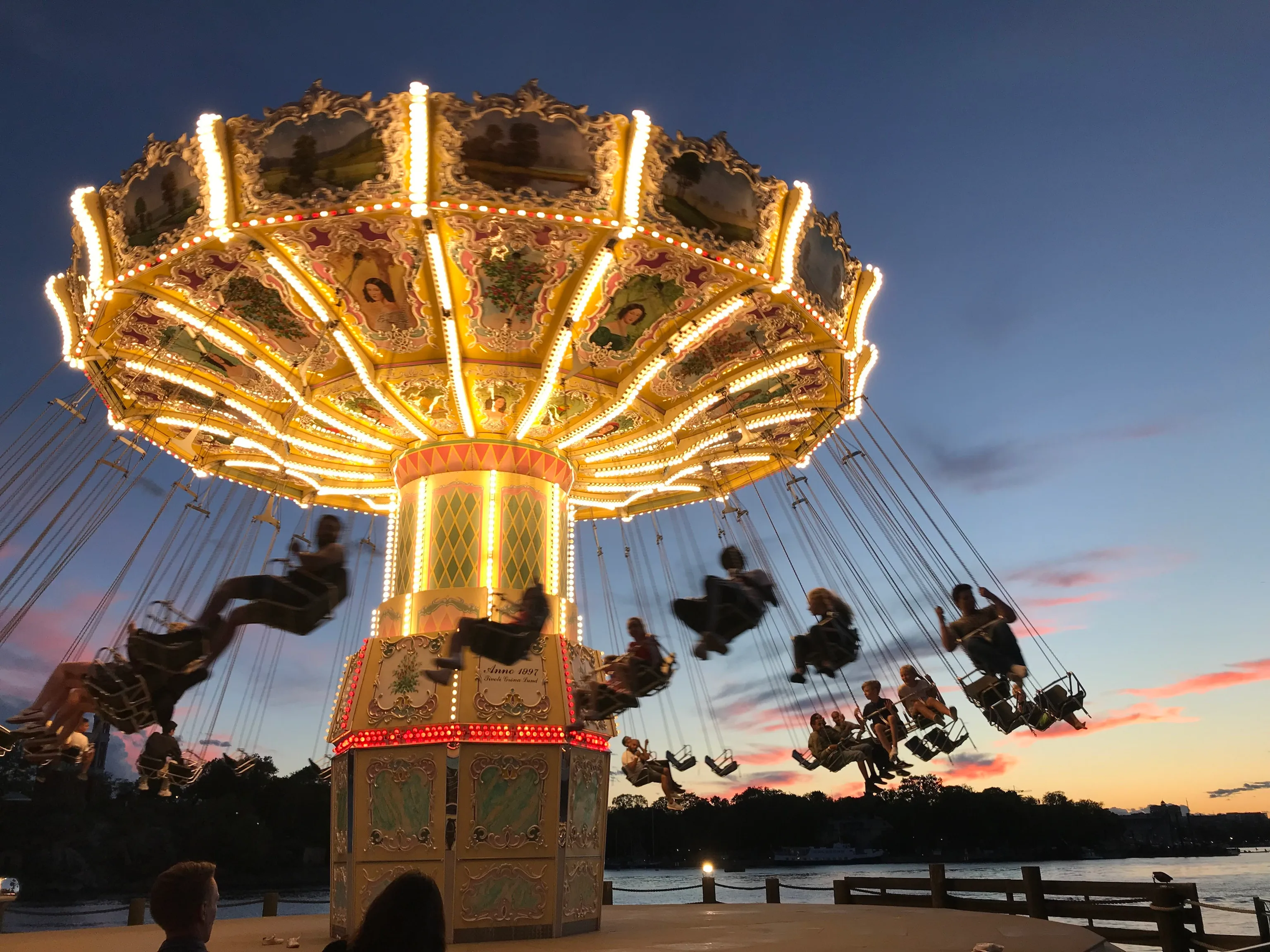 Riders swing outward on an ornate carousel chair ride glowing with golden lights, set against a twilight sky streaked with pink and blue near a waterfront.
