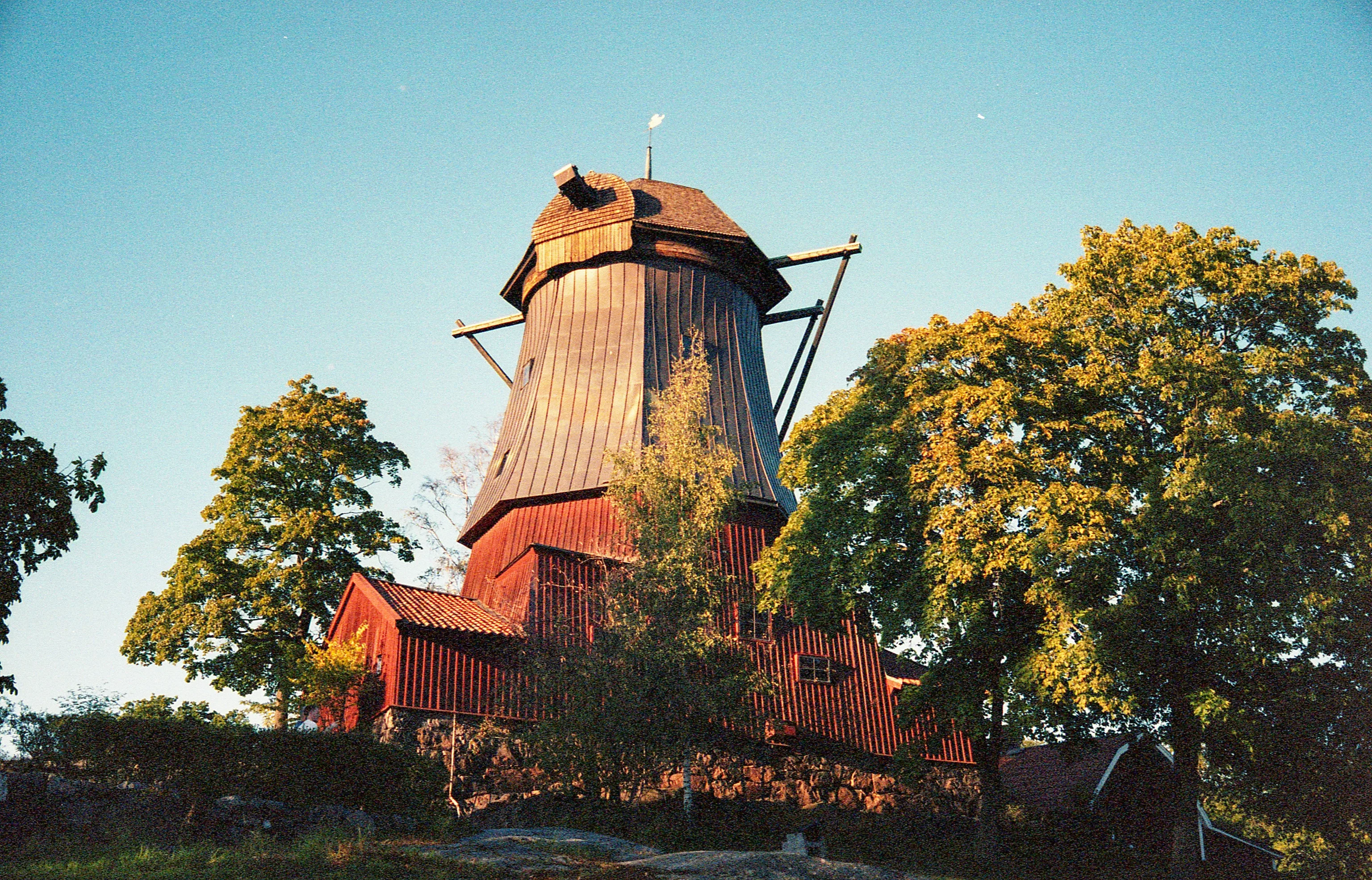 A weathered red windmill with a dark tapered roof stands among leafy green trees, viewed from below against a clear blue sky in warm golden light.