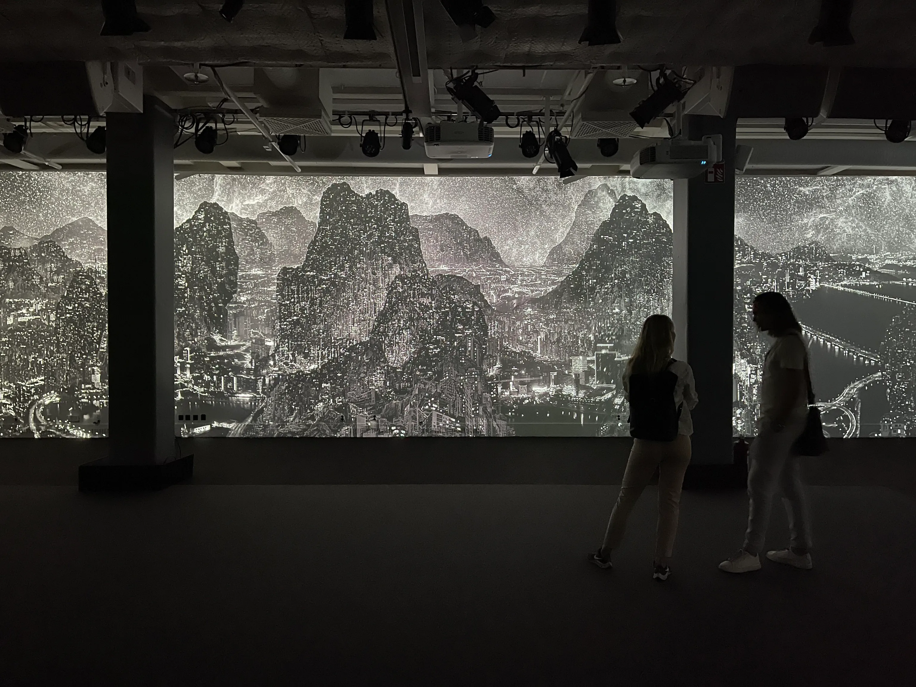 Two silhouetted visitors stand in a dark gallery watching a large glowing projection of a light-filled mountain cityscape, its white points of light spanning the wall between columns.