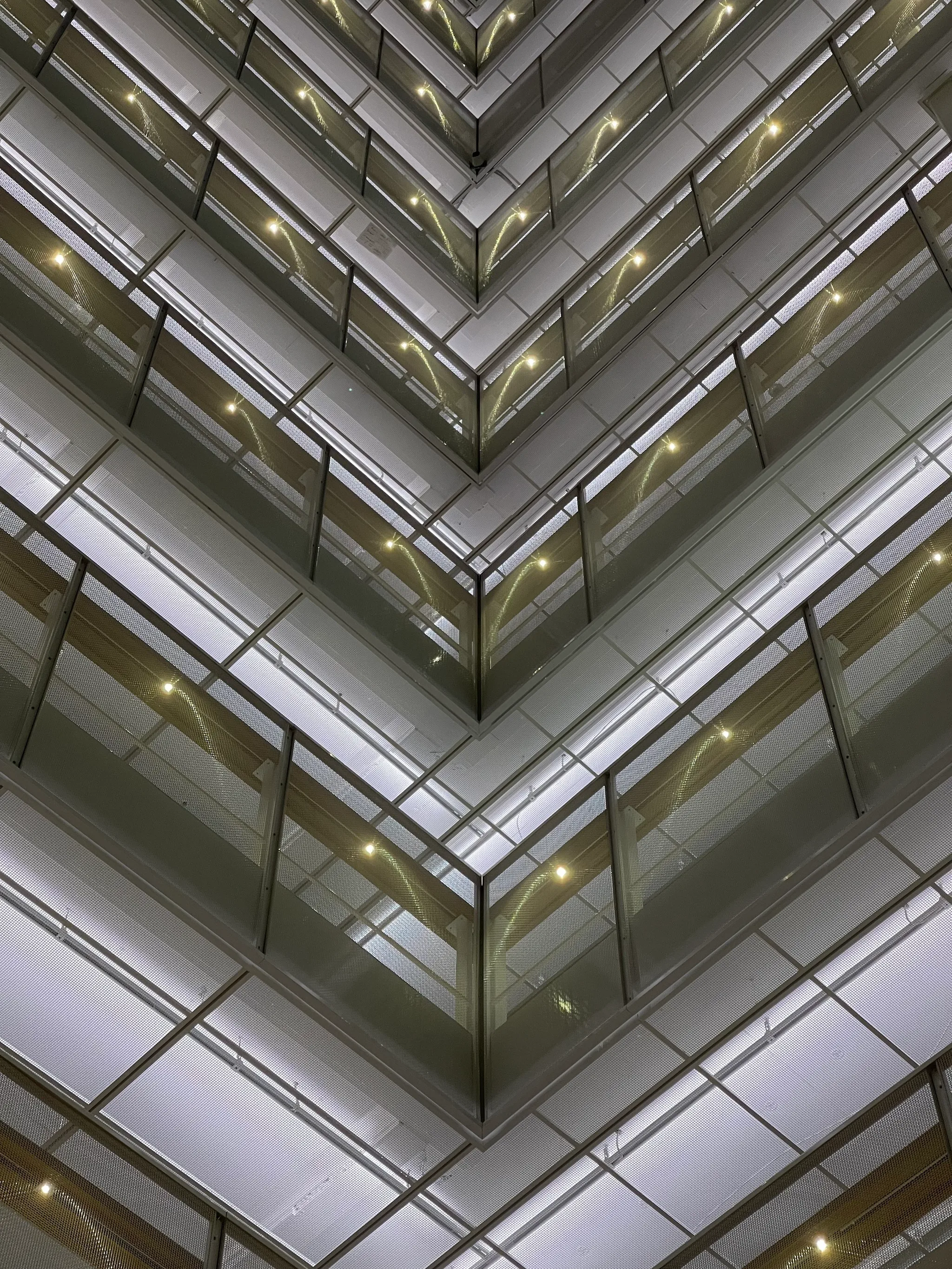 Looking up inside a multi-storey atrium, stacked glass-railed balconies form converging V-shaped tiers, each level dotted with small warm lights against pale white paneling.