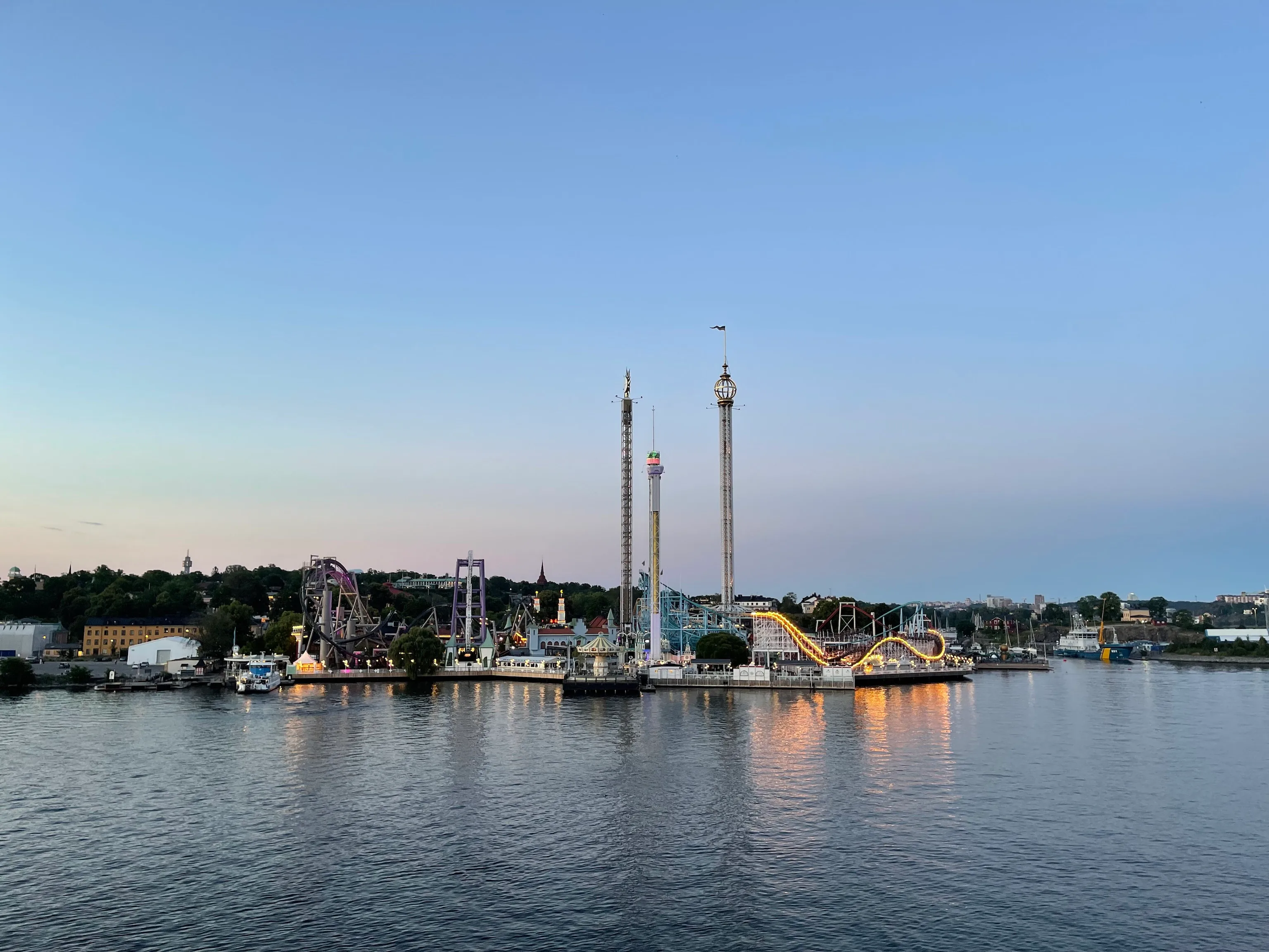 The Gröna Lund amusement park lines a calm waterfront at dusk, its tall drop towers and warmly lit roller coaster glowing against a soft blue sky.