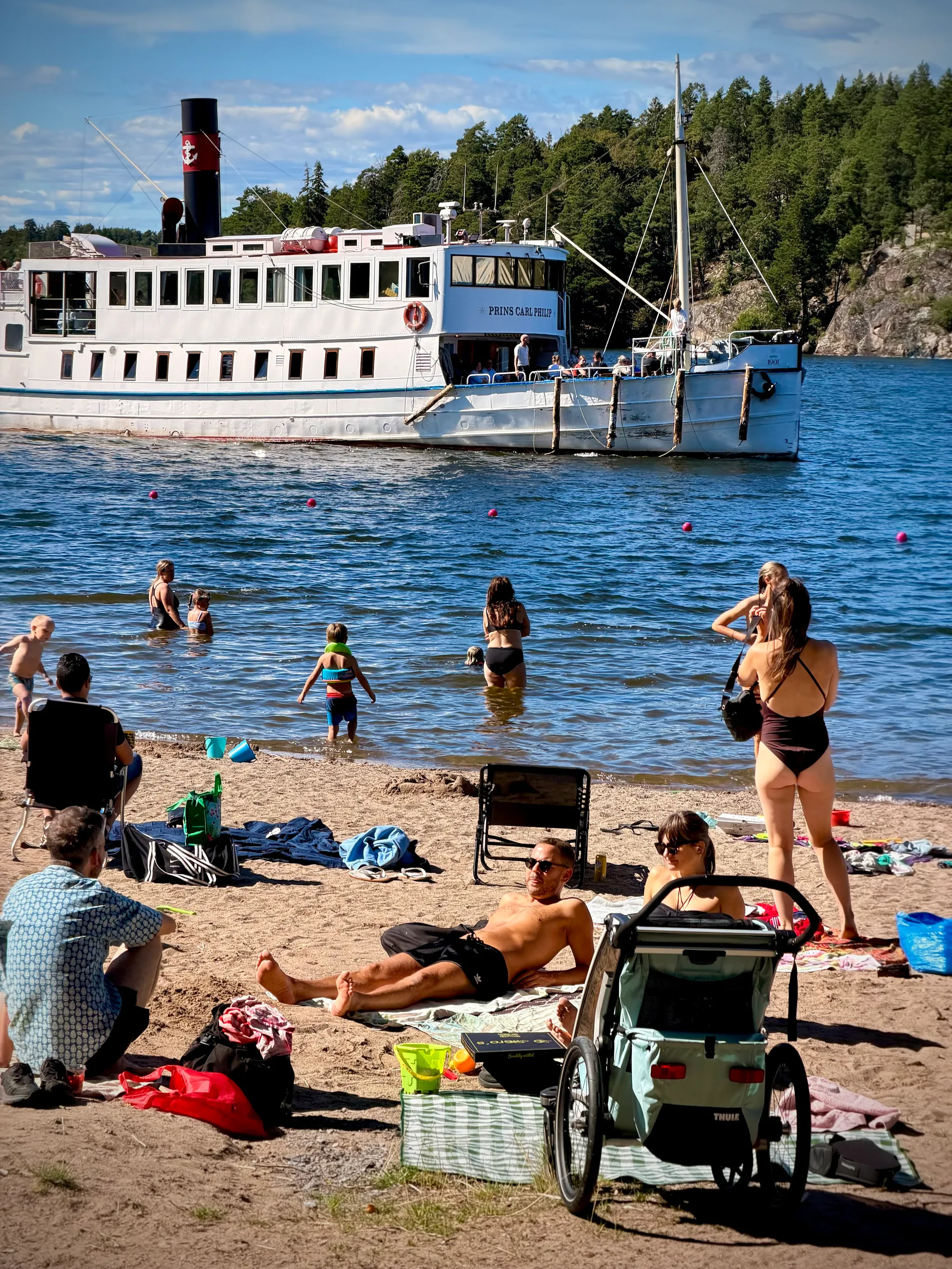 Beachgoers sunbathe on towels and wade into the water as a white vintage steamship named Prins Carl Philip glides past a forested shoreline under a bright blue sky.