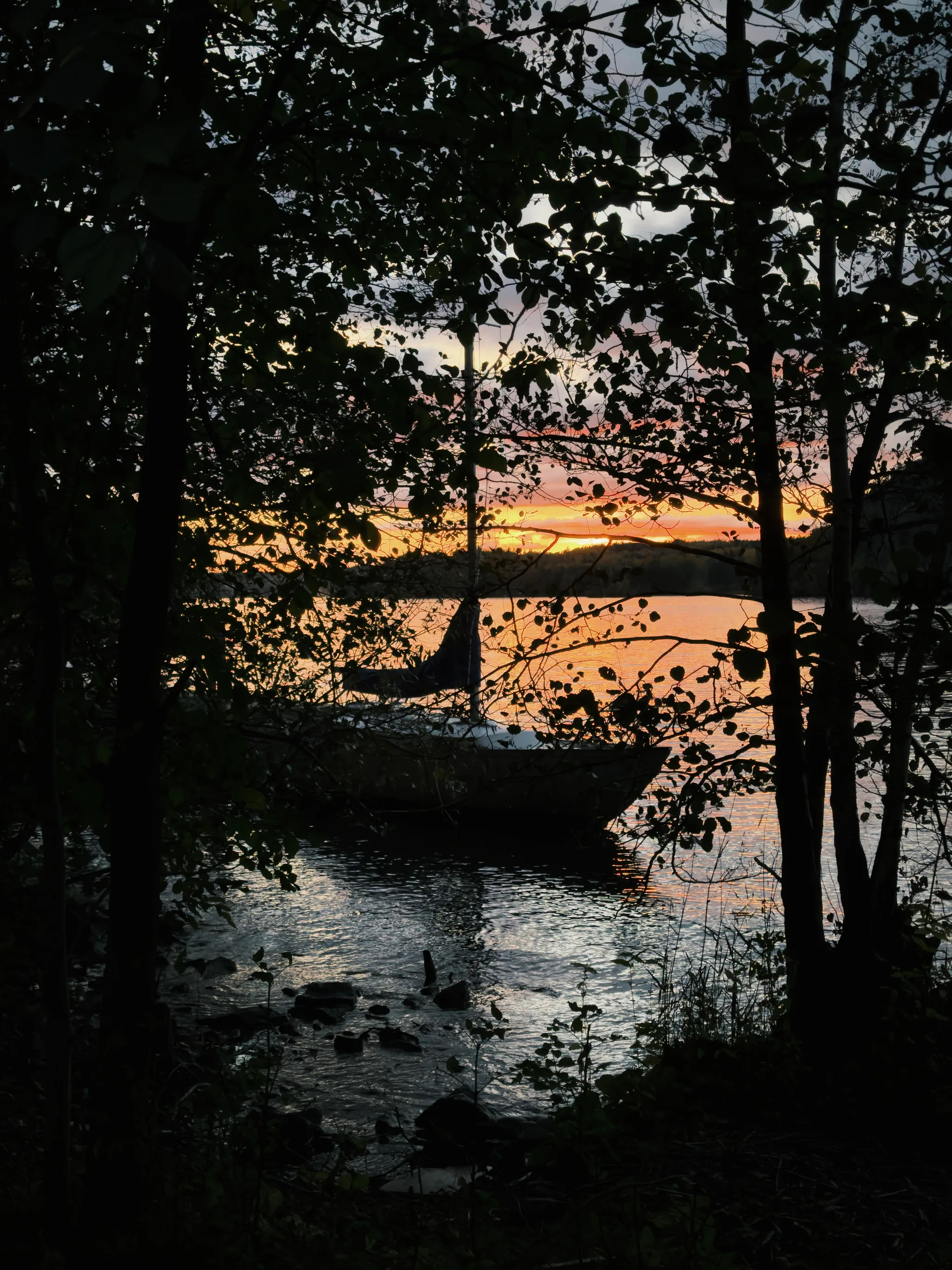 A sailboat rests on calm water at dusk, framed by dark silhouetted trees, with a glowing orange and yellow sunset reflected across the lake's surface.