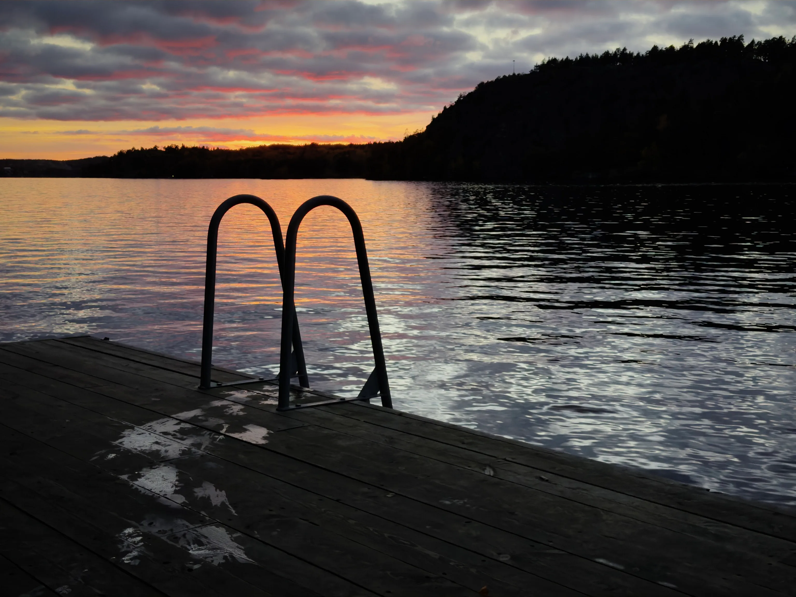 A metal swim ladder stands at the end of a wooden dock overlooking a calm lake at sunset, with remnants of snow on the planks and a colorful orange and pink sky above dark trees.