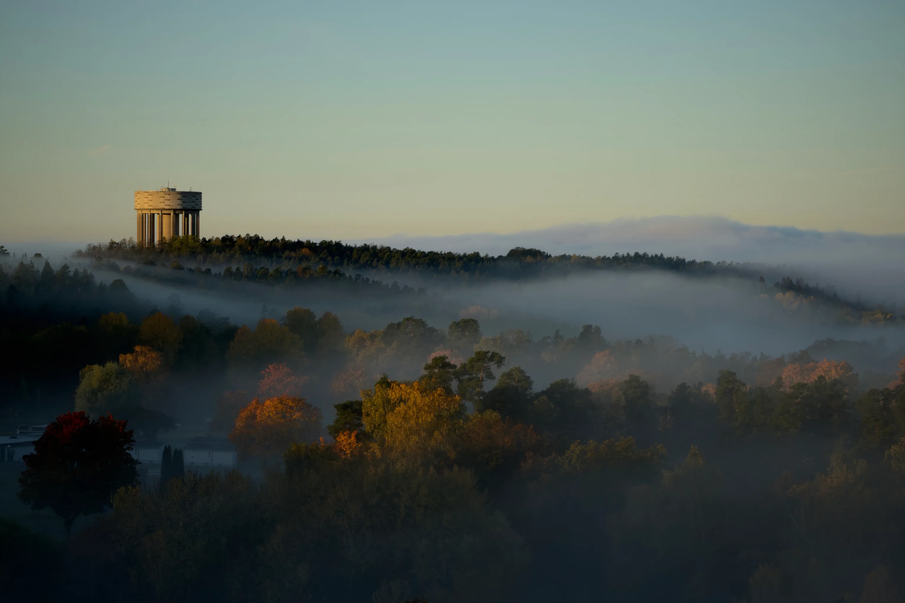 Low morning fog rolls over autumn treetops while a cylindrical water tower rises from a misty hilltop beneath a soft pale green and blue sky.