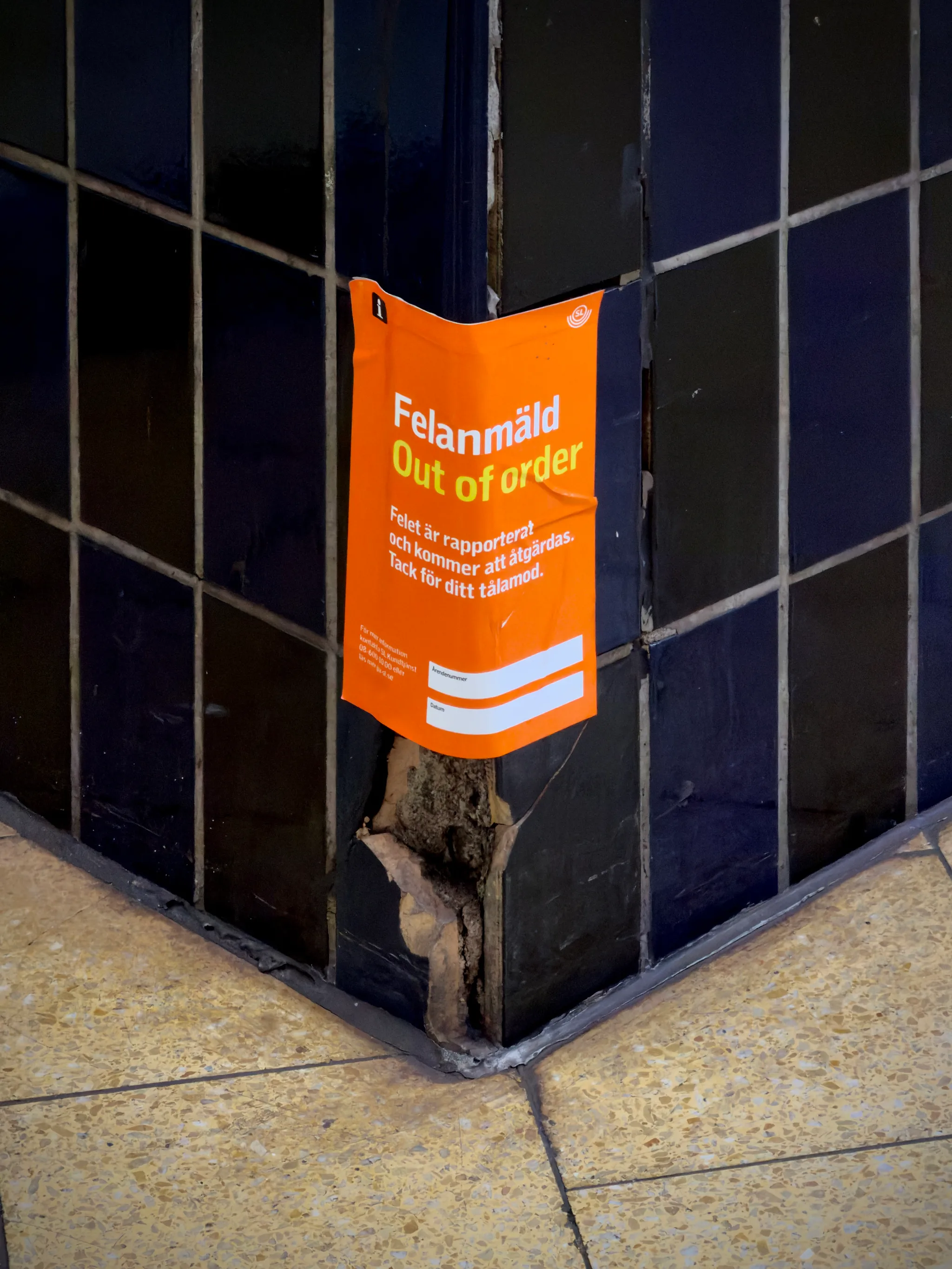 A bright orange flyer wrapped around the corner of a glossy black tiled pillar is peeling at the bottom, revealing torn paper against dark blue squares and beige floor tiles.